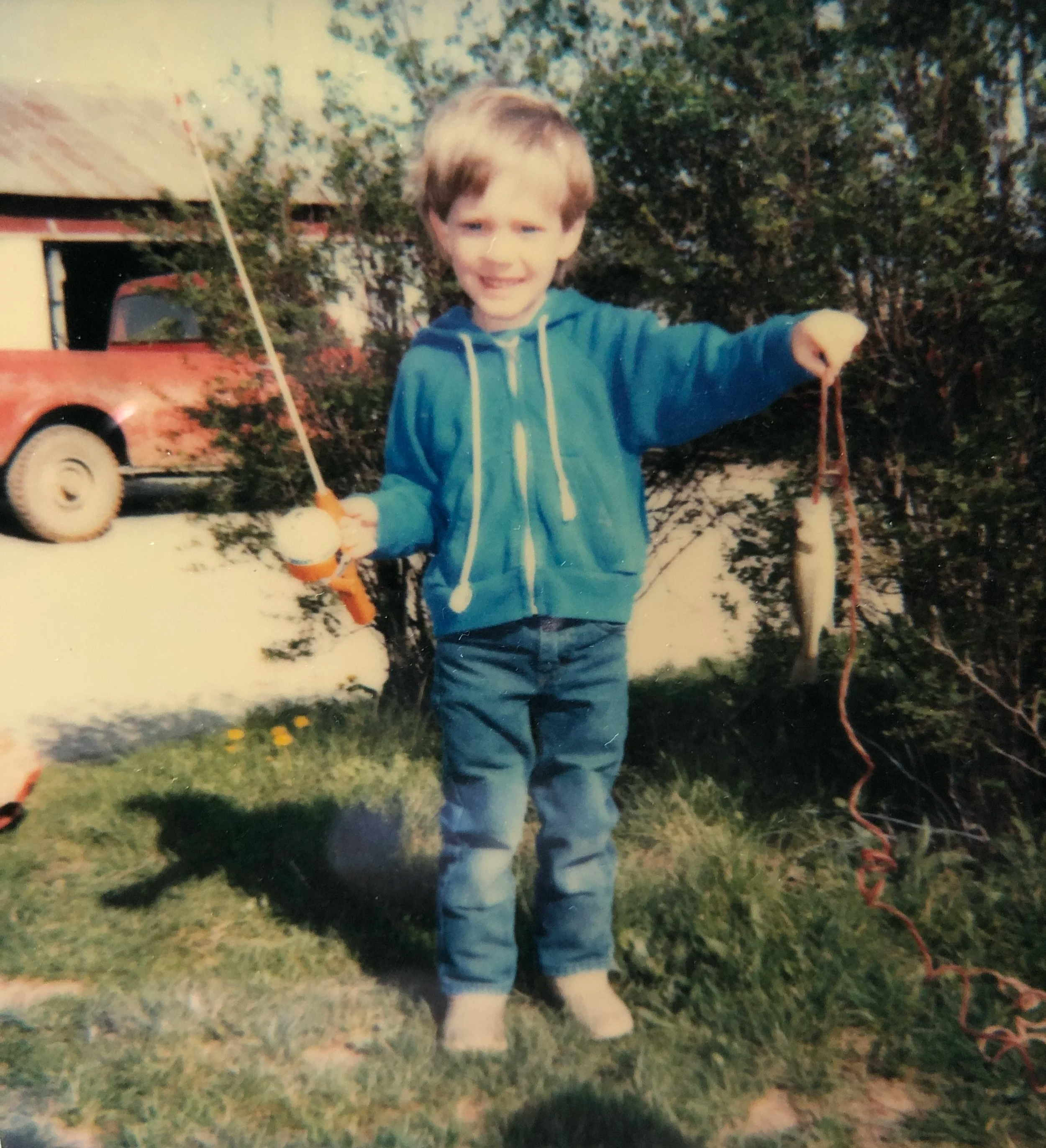 A young boy wearing a blue hoodie and jeans holding a toy boat with a string, smiling outdoors in front of trees and a house, with a red vehicle nearby.