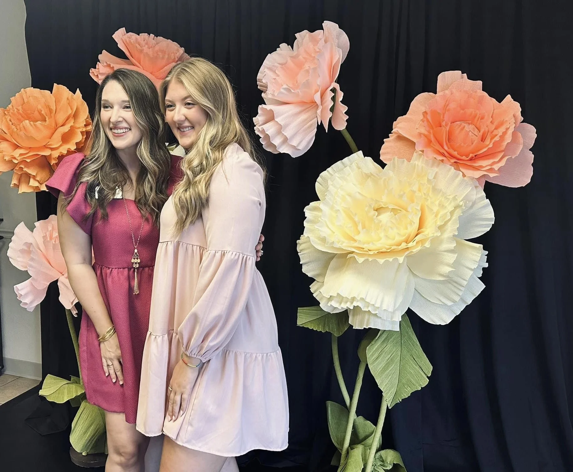 Two women smiling and standing close together in front of a large display of flowers, with a dark background.