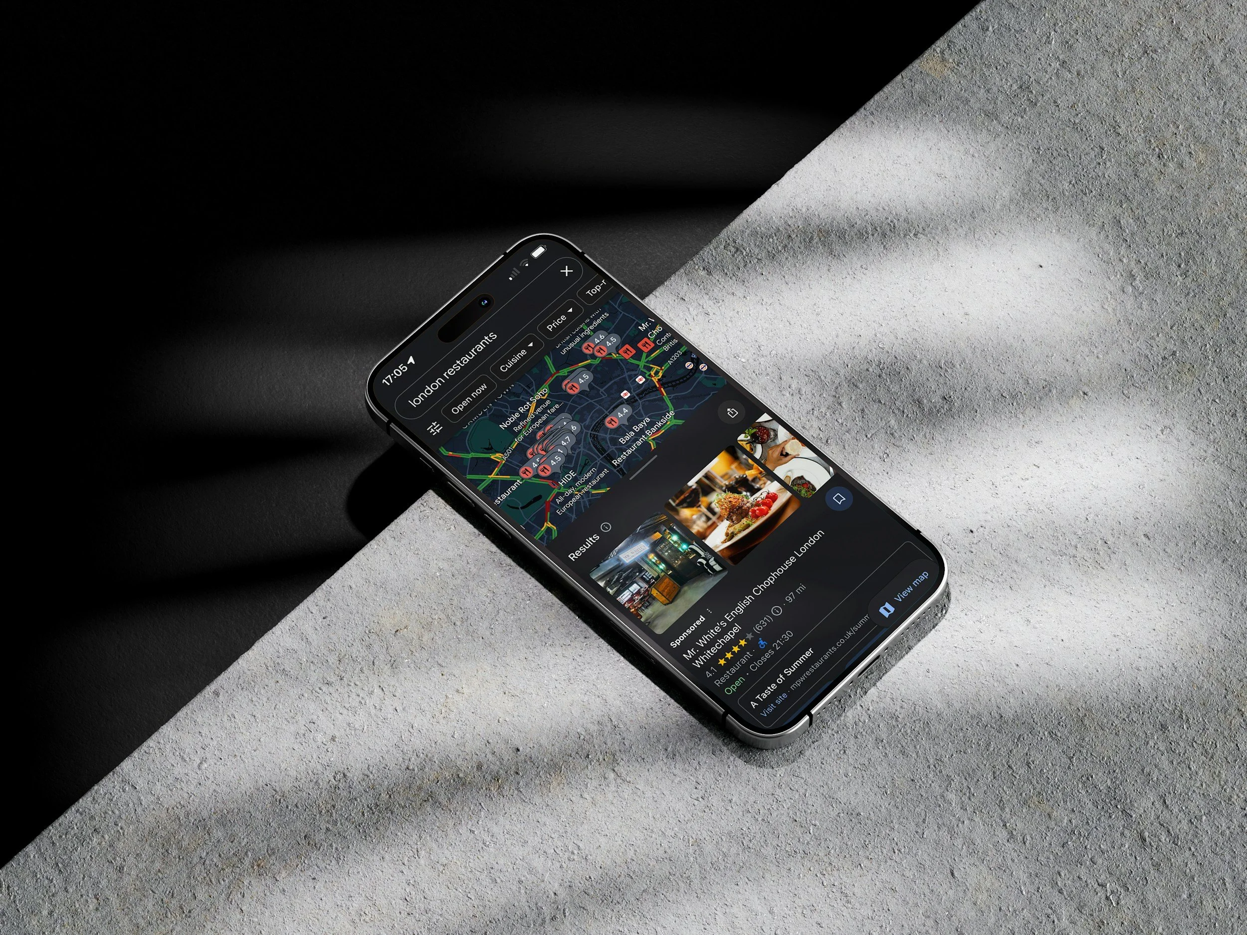 Smartphone displaying a map and restaurant search results on a dark background, placed on a partially lit concrete surface.