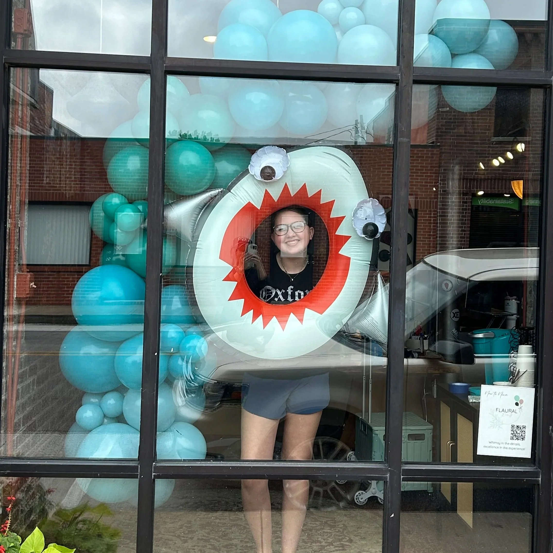 Woman smiling in a window decorated with a large inflatable shark and balloons in shades of blue, with her reflection and interior of the shop visible.