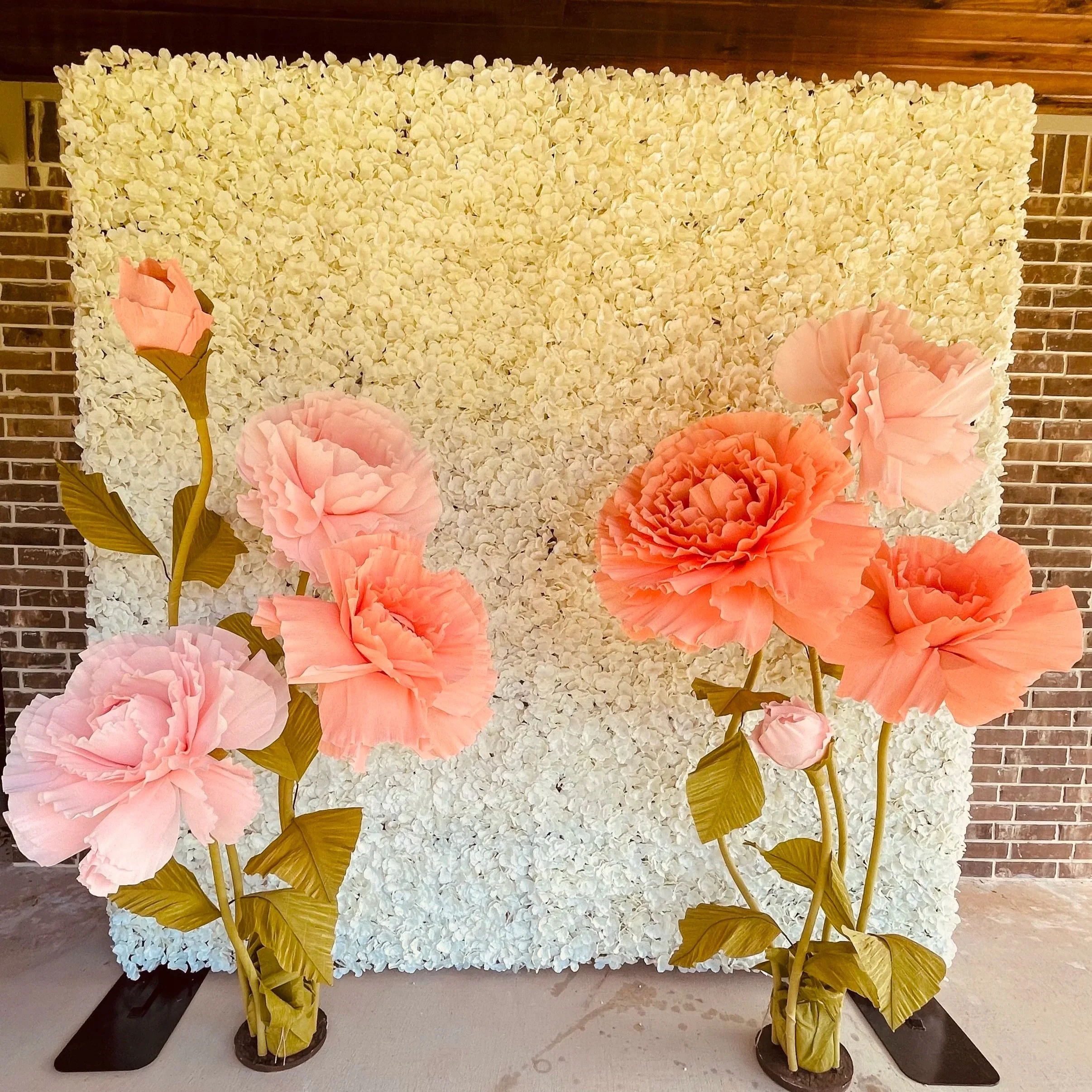 Decorative backdrop made of white flowers with large pink flowers and green leaves in front, set against a brick wall.