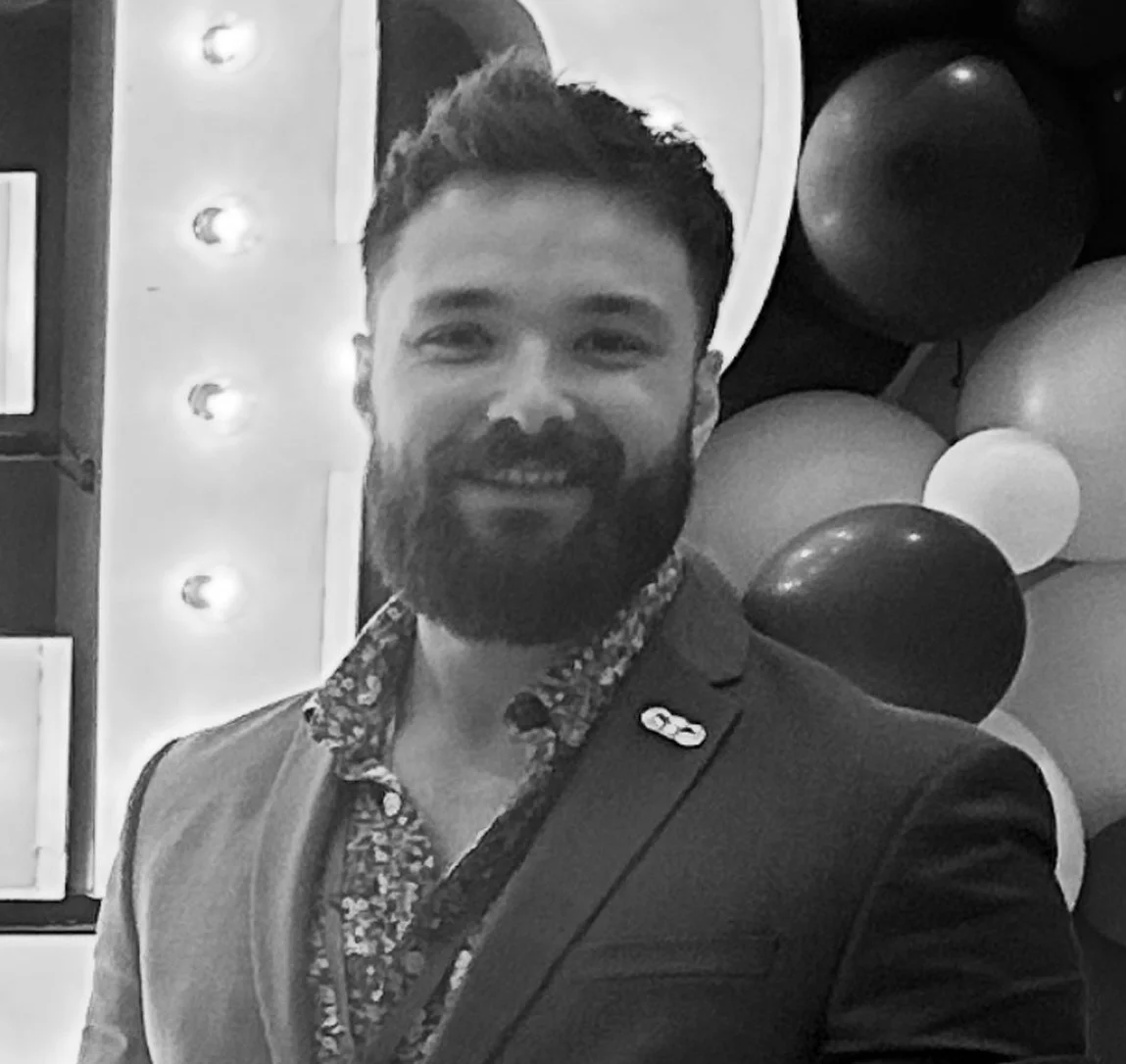 A smiling man with a beard and mustache, wearing a blazer and a patterned shirt, standing in front of a backdrop with balloons and lights.