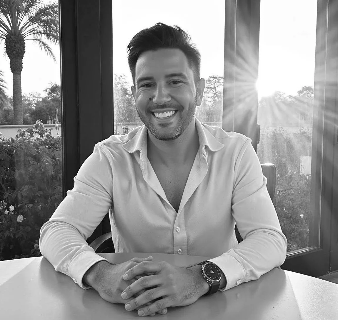 A smiling man with short dark hair and a beard, wearing a light-colored button-up shirt and a watch, sitting at a table with his hands clasped. He is indoors near a window with a view of trees and plants outside, with sunlight streaming in.