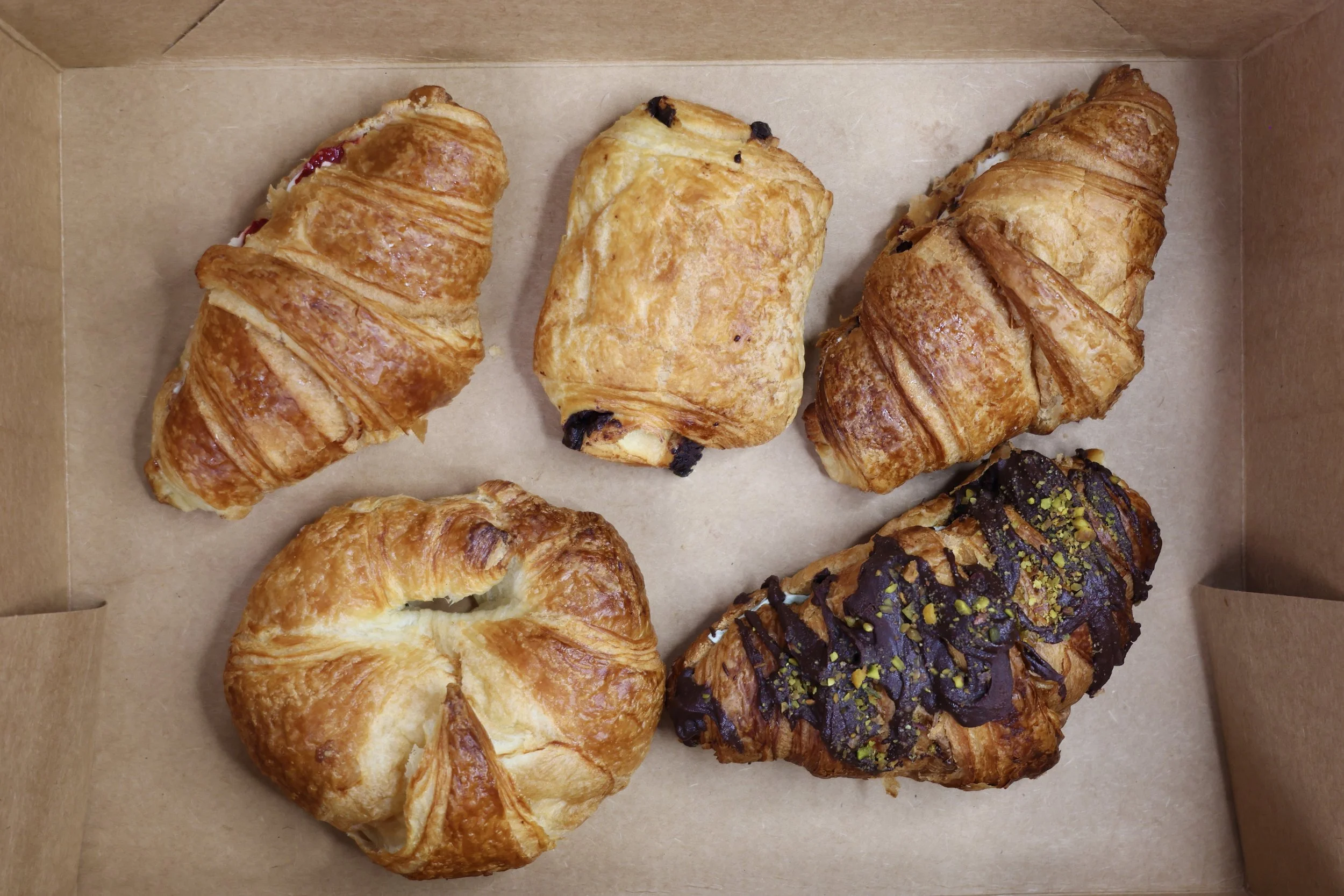 Six assorted pastries in a cardboard box, including croissants, a Danish pastry, and a chocolate-covered pastry with pistachio topping.