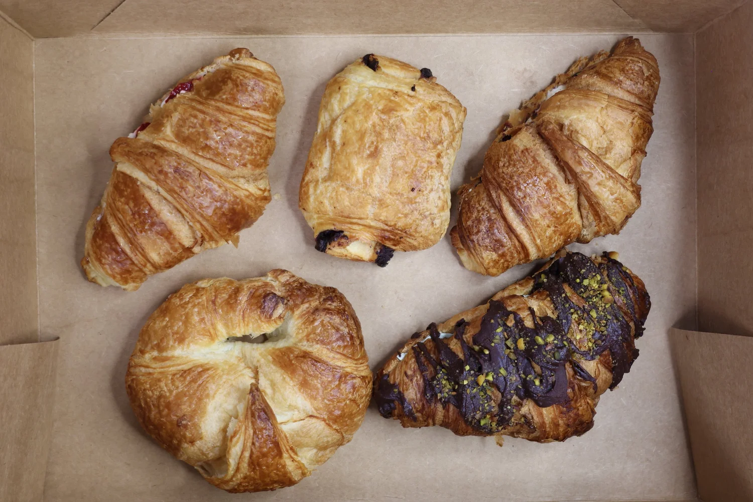 A kraft box of croissants, danishes and a chocolate pistachio pastry