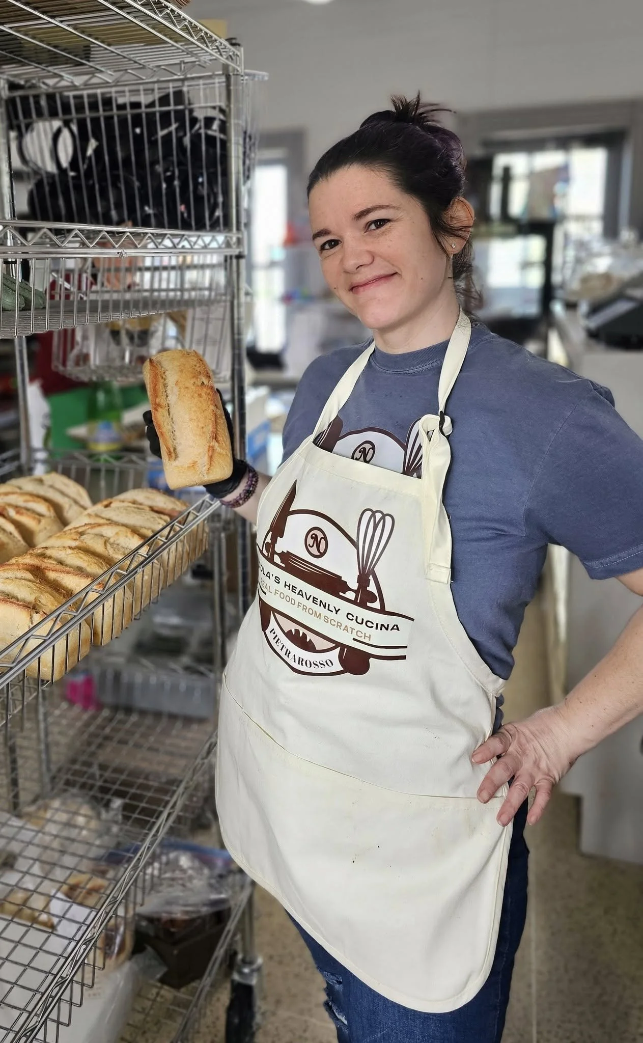 A woman standing in a kitchen or bakery, wearing a white apron with a logo, holding a toasted sandwich in front of a metal rack filled with more sandwiches. She has dark hair tied up and is smiling at the camera.