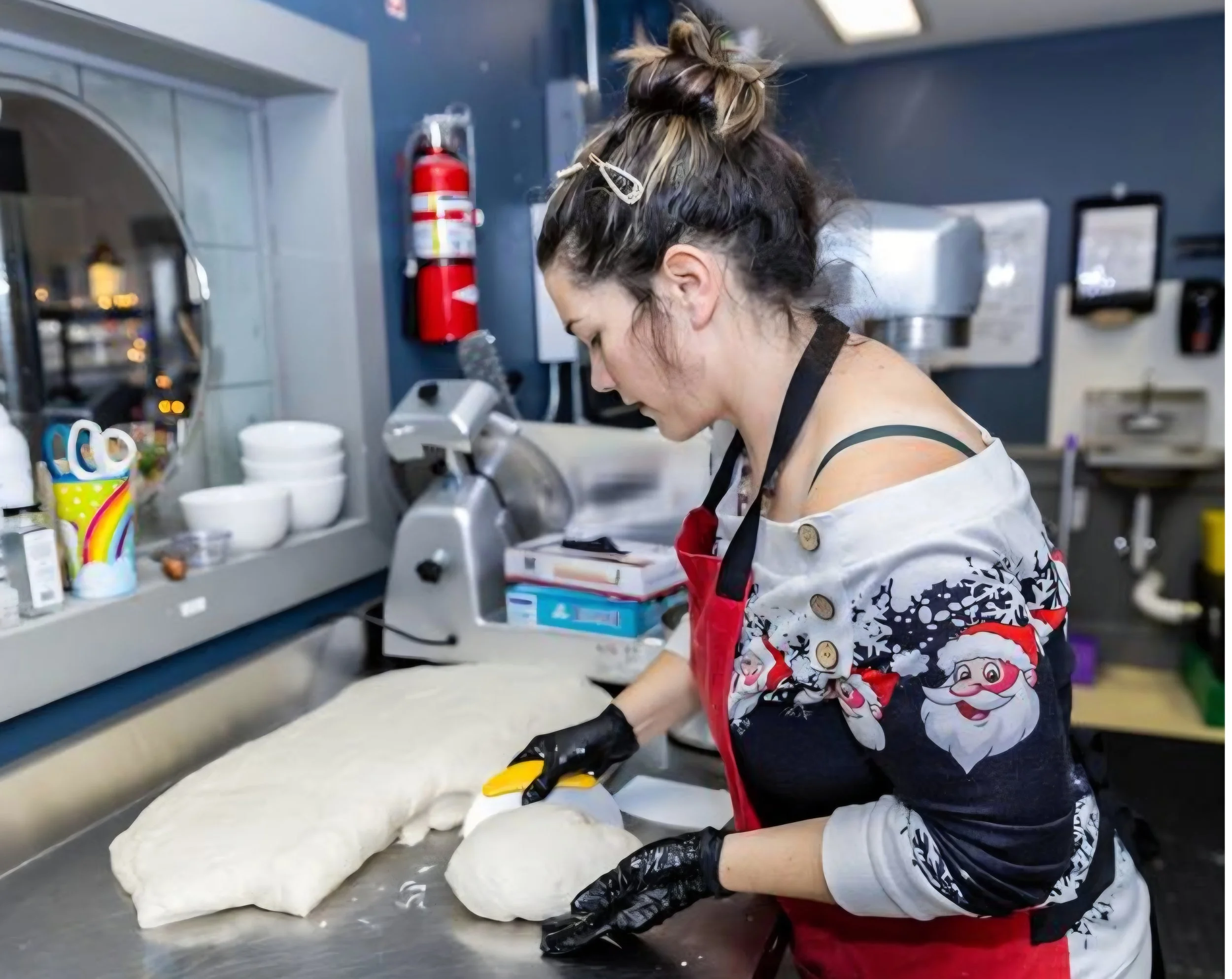 A person with hair tied up working in a commercial kitchen, wearing a Christmas-themed apron, preparing dough with a rolling pin on a gray countertop.