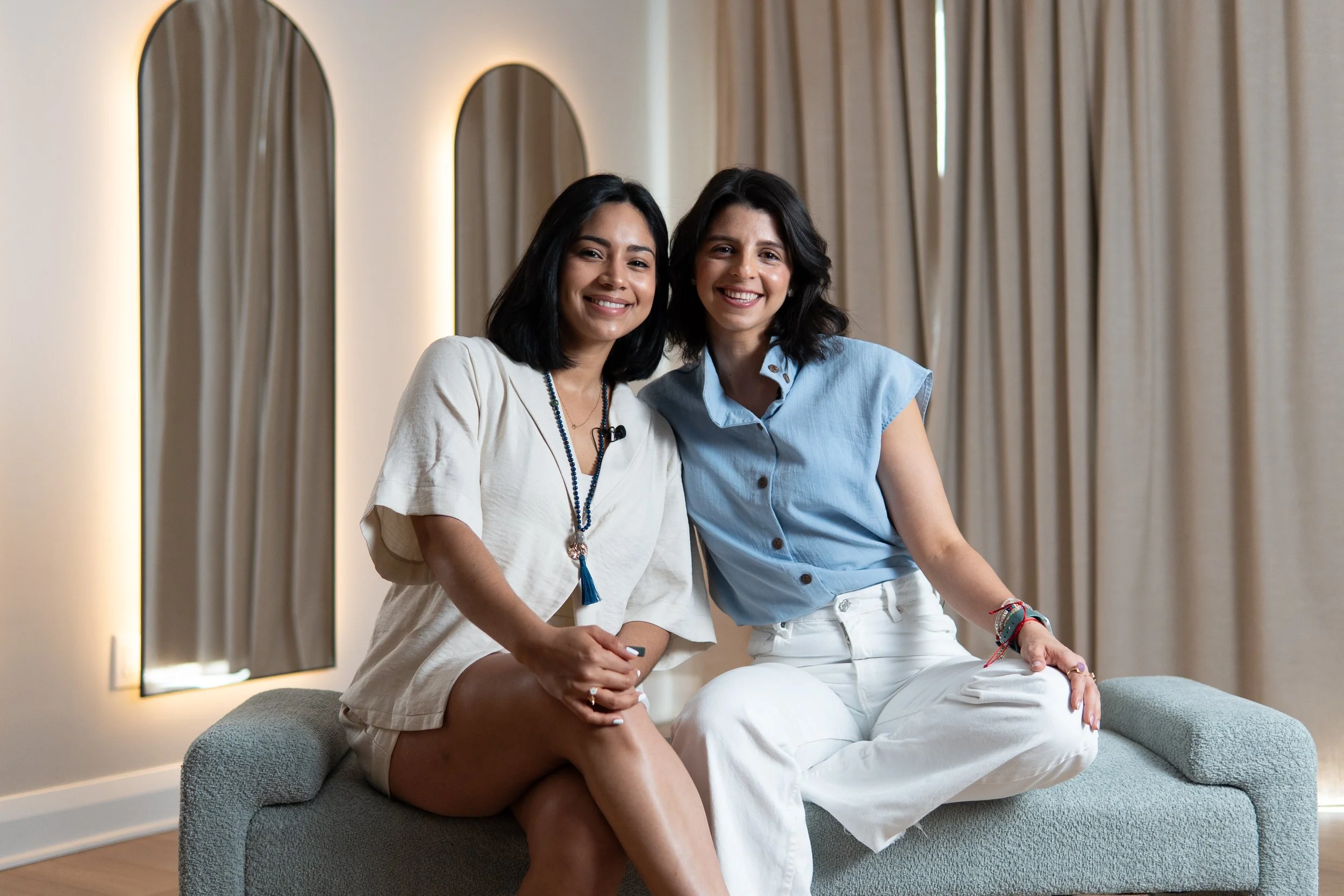 Two women sitting on a gray couch in front of beige curtains, smiling at the camera. One woman is wearing a white shirt and the other is wearing a light blue shirt.