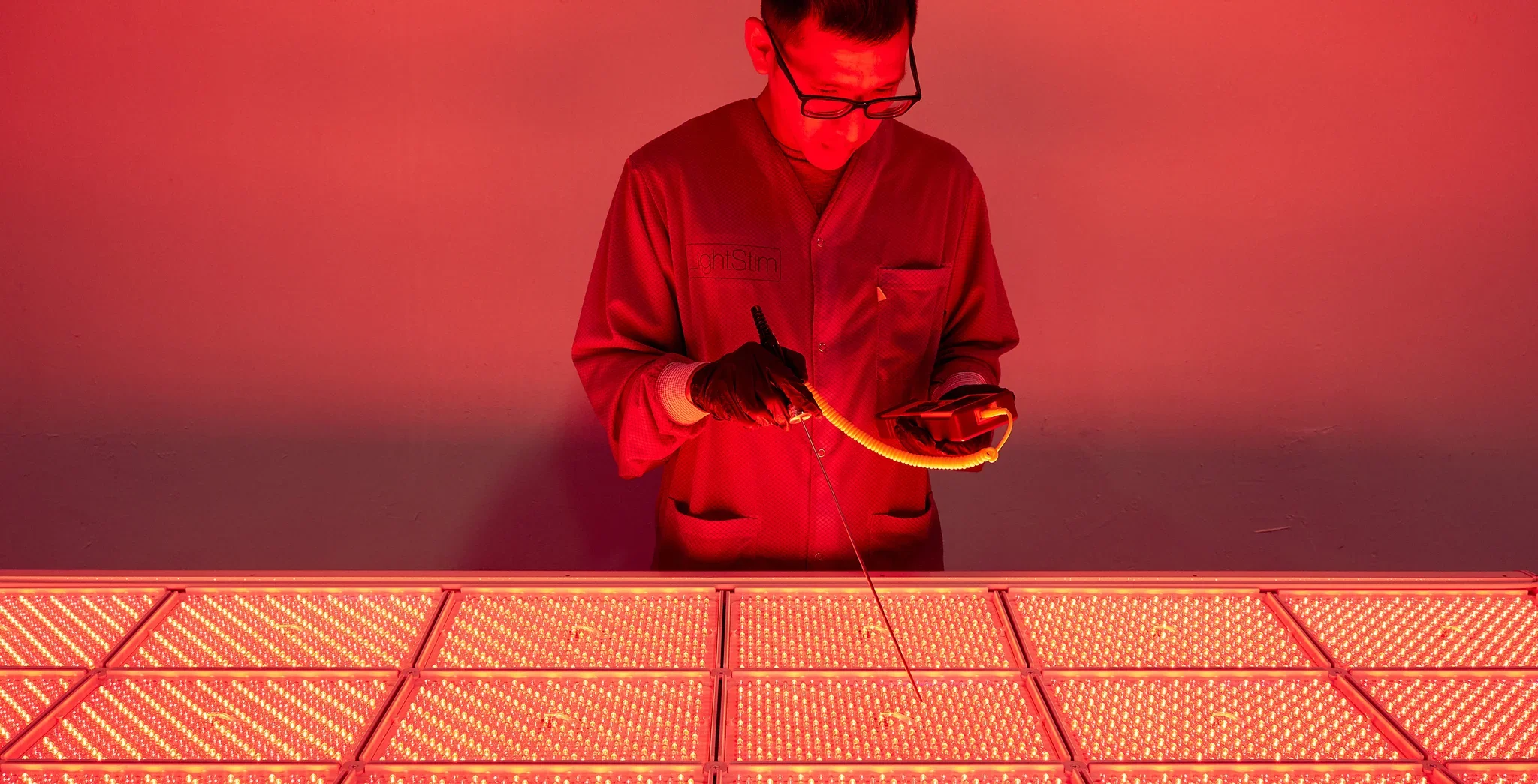 A LightStim+ ProPanel LED light therapy device emitting red light, positioned above a brown upholstered couch next to a window with green foliage outside.