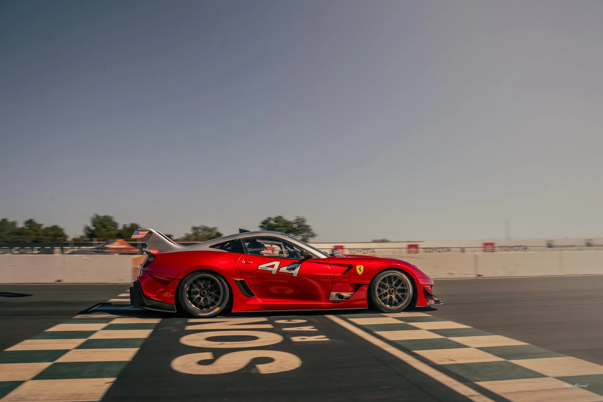 A red Ferrari race car crossing the finish line on a race track during daytime with a checkered flag pattern on the ground and some trees and barriers in the background.