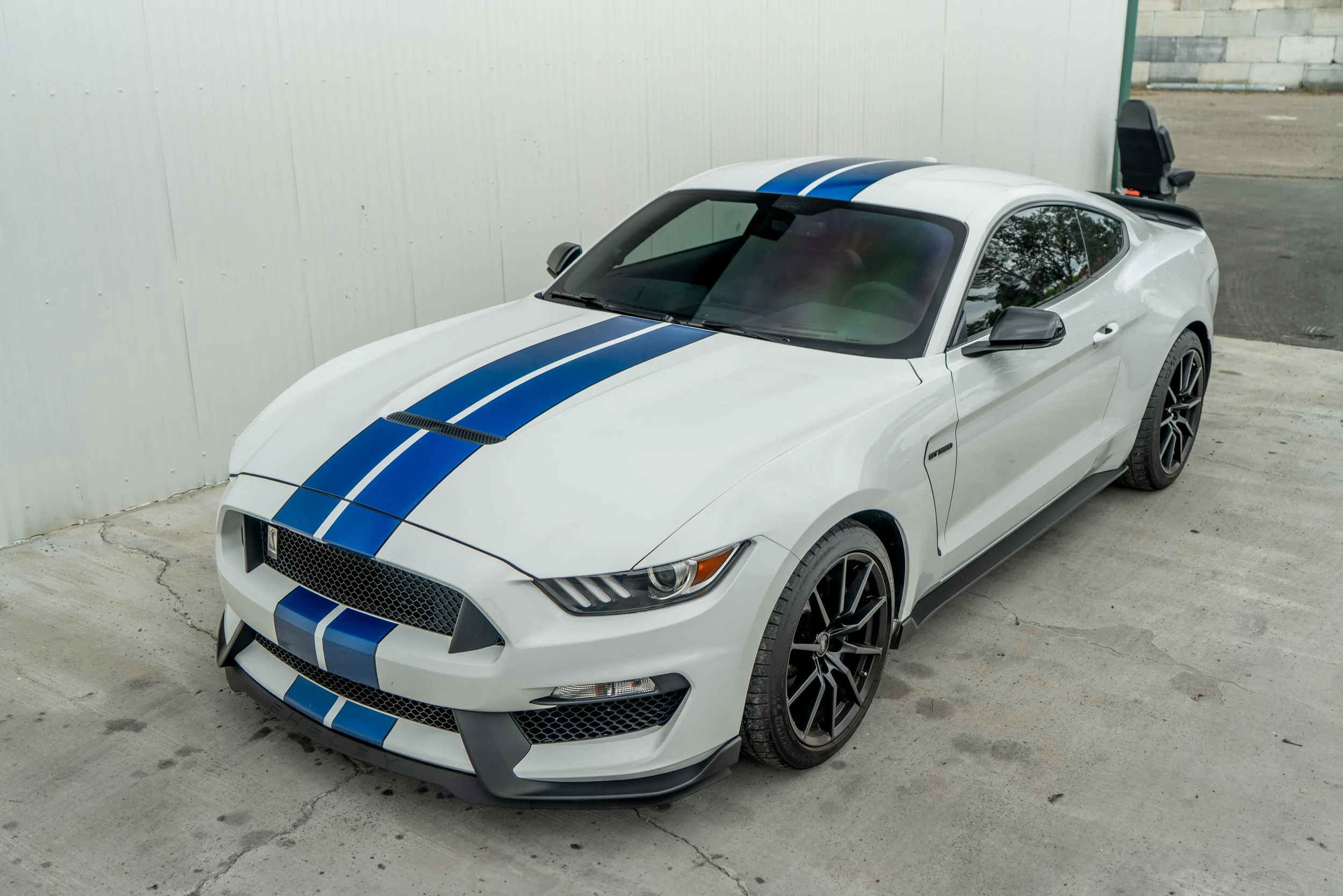 White sports car with blue racing stripes parked on concrete surface near a metal wall.