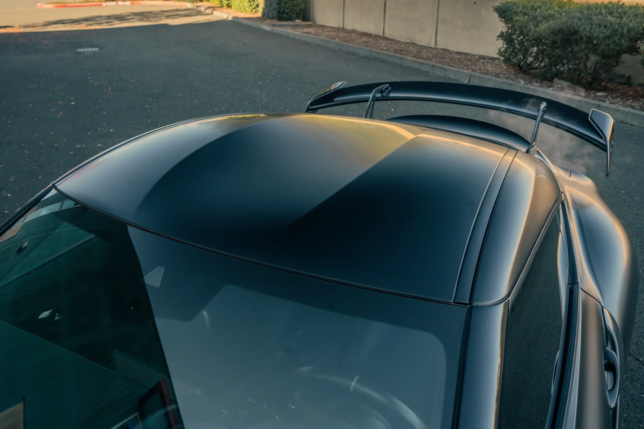 Top view of a dark-colored sports car parked on an asphalt road, with a large rear wing and a sleek, shiny surface reflecting sunlight.