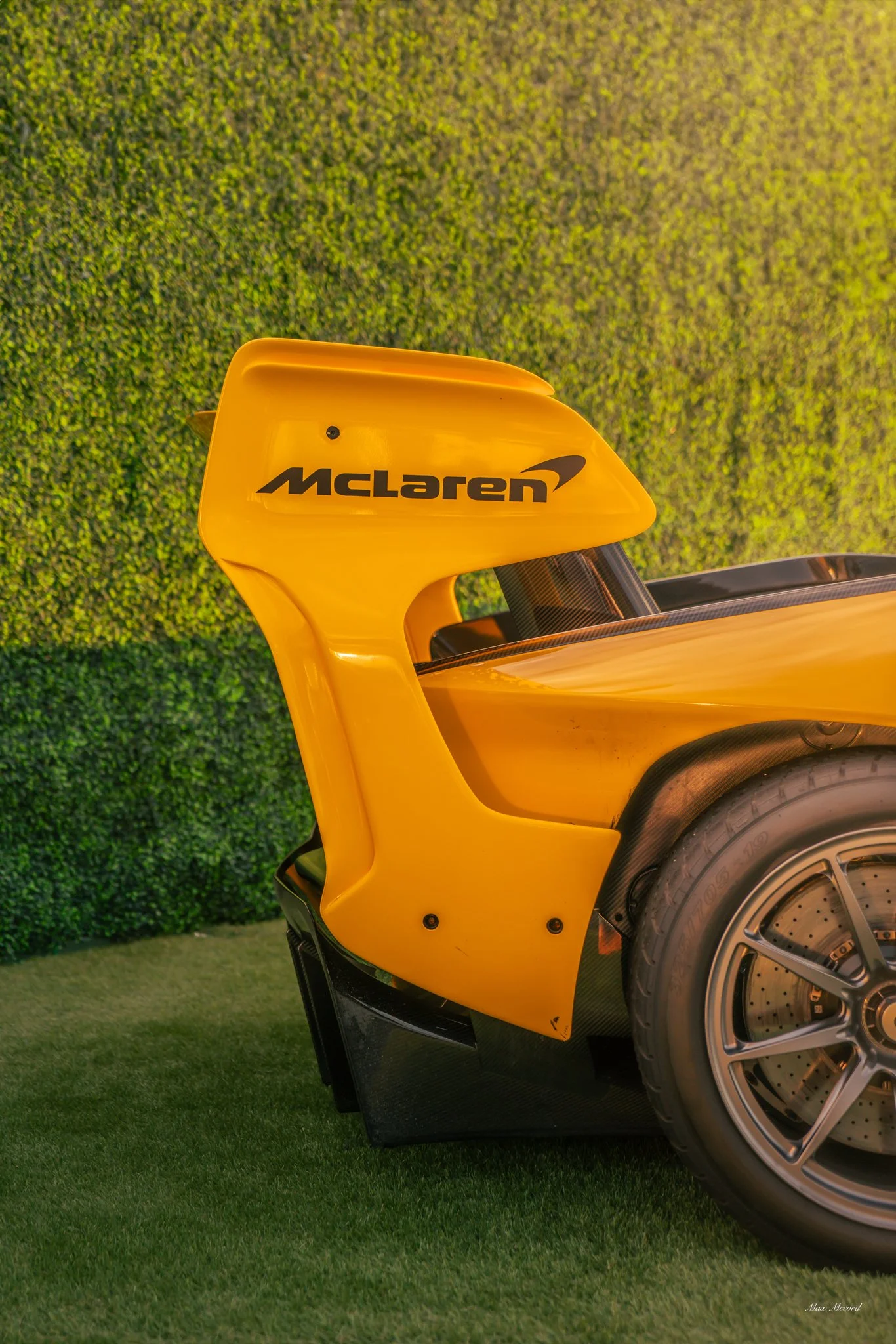 Close-up of the rear wing of a yellow McLaren F1 race car, with the McLaren logo visible on the wing, against a green hedge background.