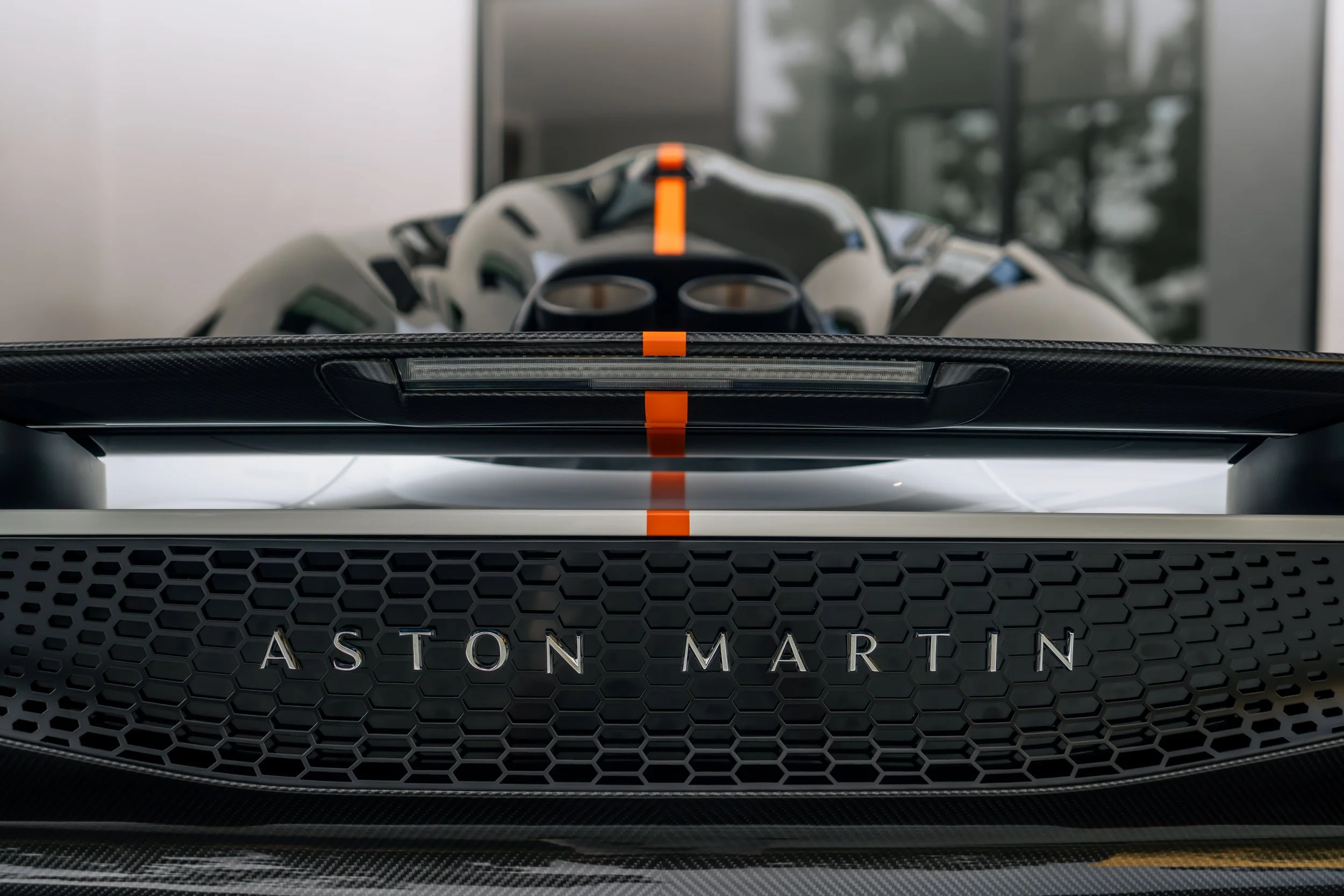 Close-up of the rear of an Aston Martin Valkyrie hypercar with the car's logo displayed on a honeycomb grille, a helmet with a visor resting on the car's rear, and a modern building in the background.