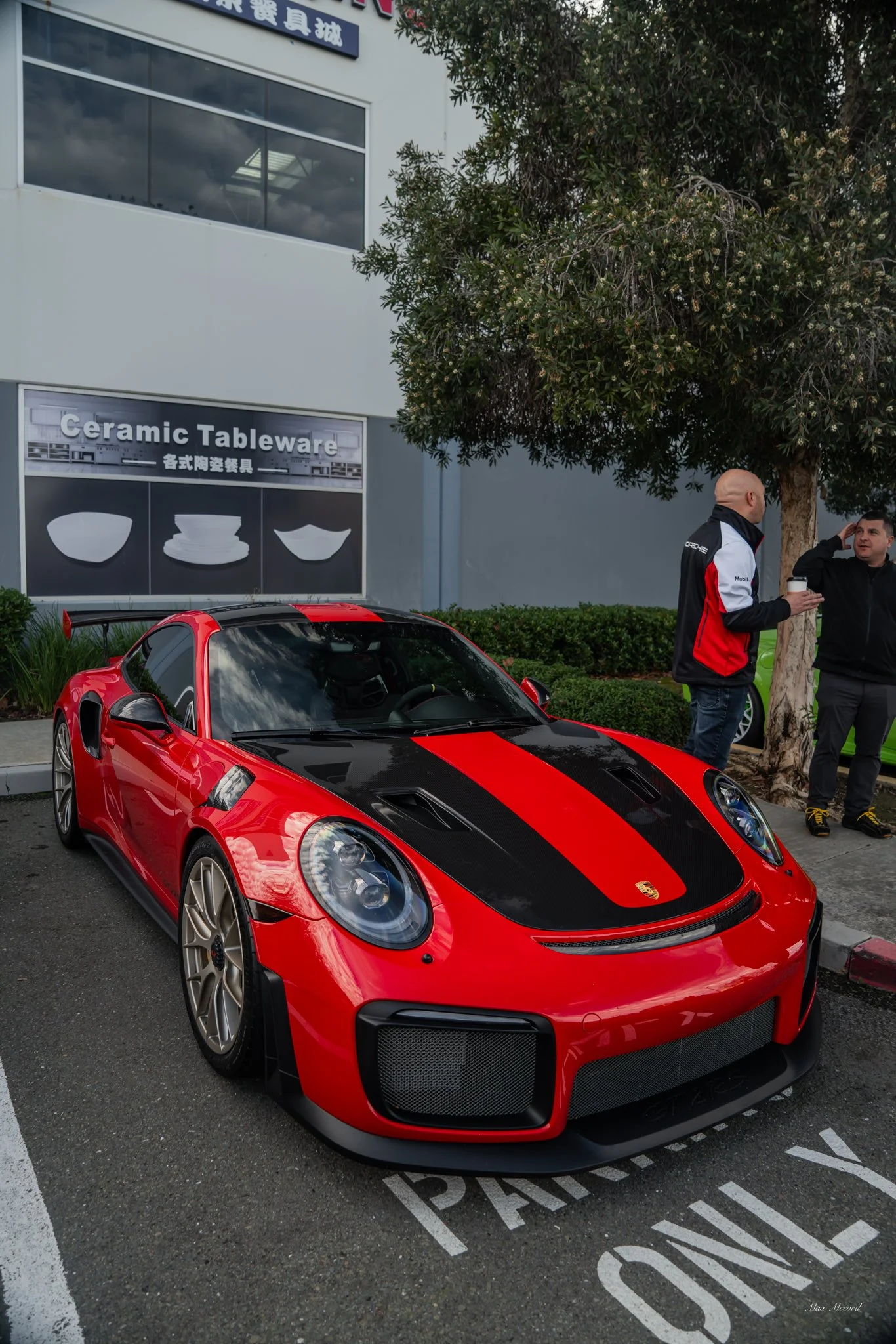 A red Porsche sports car parked in a parking space with the word 'PASS ONLY' painted on the ground. Two men are standing near the car, engaged in conversation, one holding a coffee cup. Behind them is a business with a sign that reads 'Ceramic Tablew