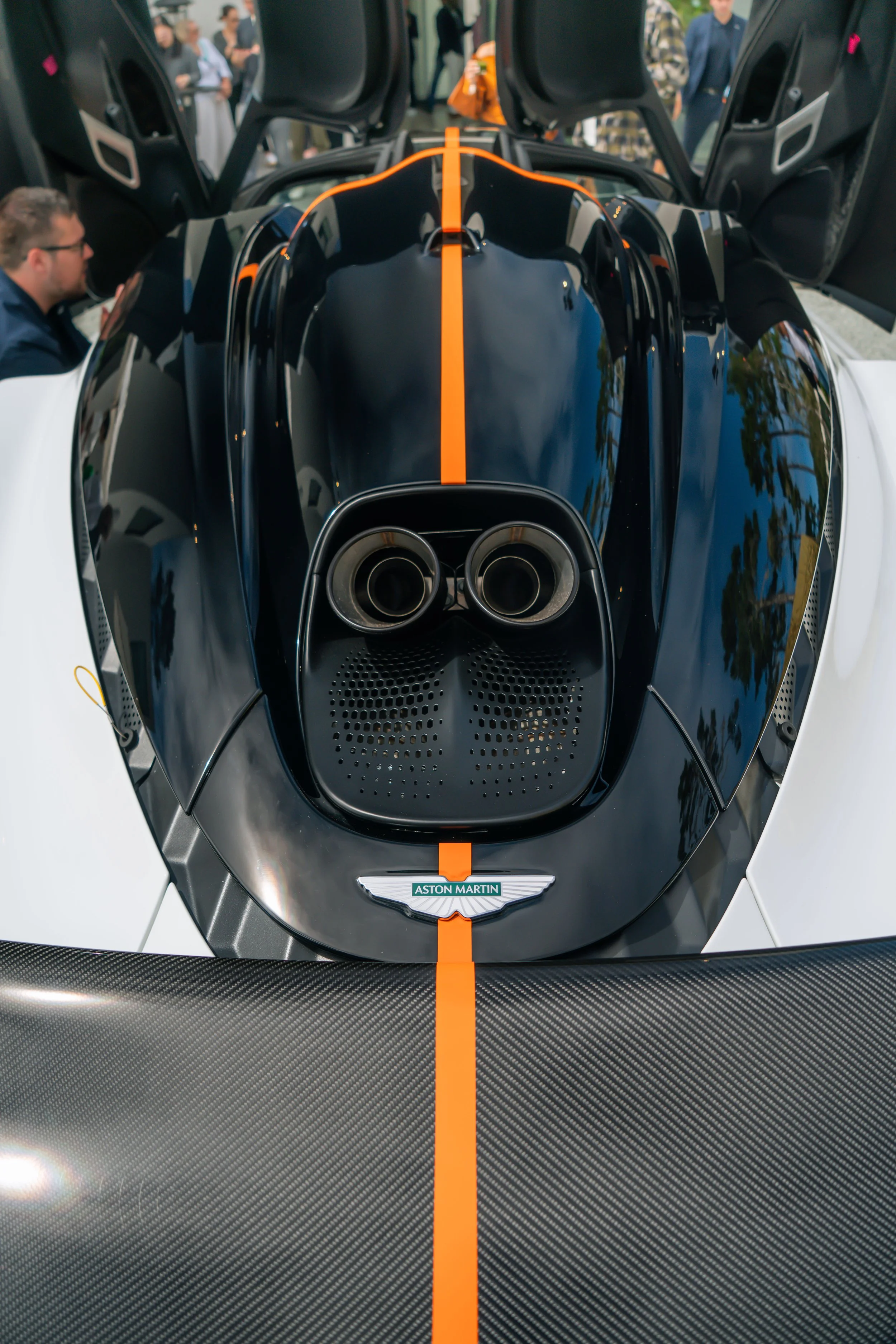 Front view of a black and white Aston Martin racing car with an orange stripe, open gull-wing doors, and visible exhaust pipes, surrounded by people.