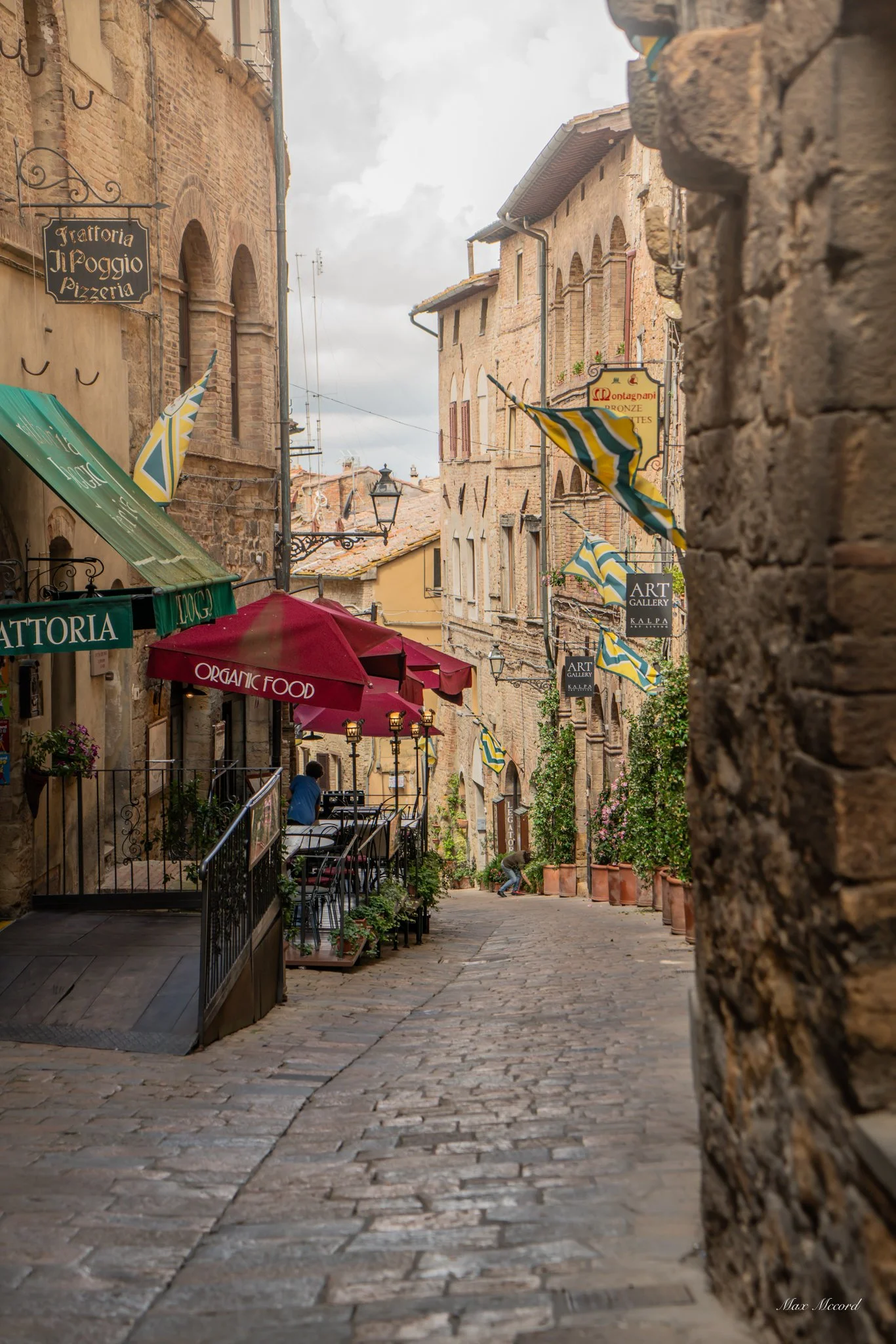 A narrow cobblestone street in Italy with outdoor seating, colorful umbrellas, and buildings with brick facades, flags, and signs for art galleries and cafes.