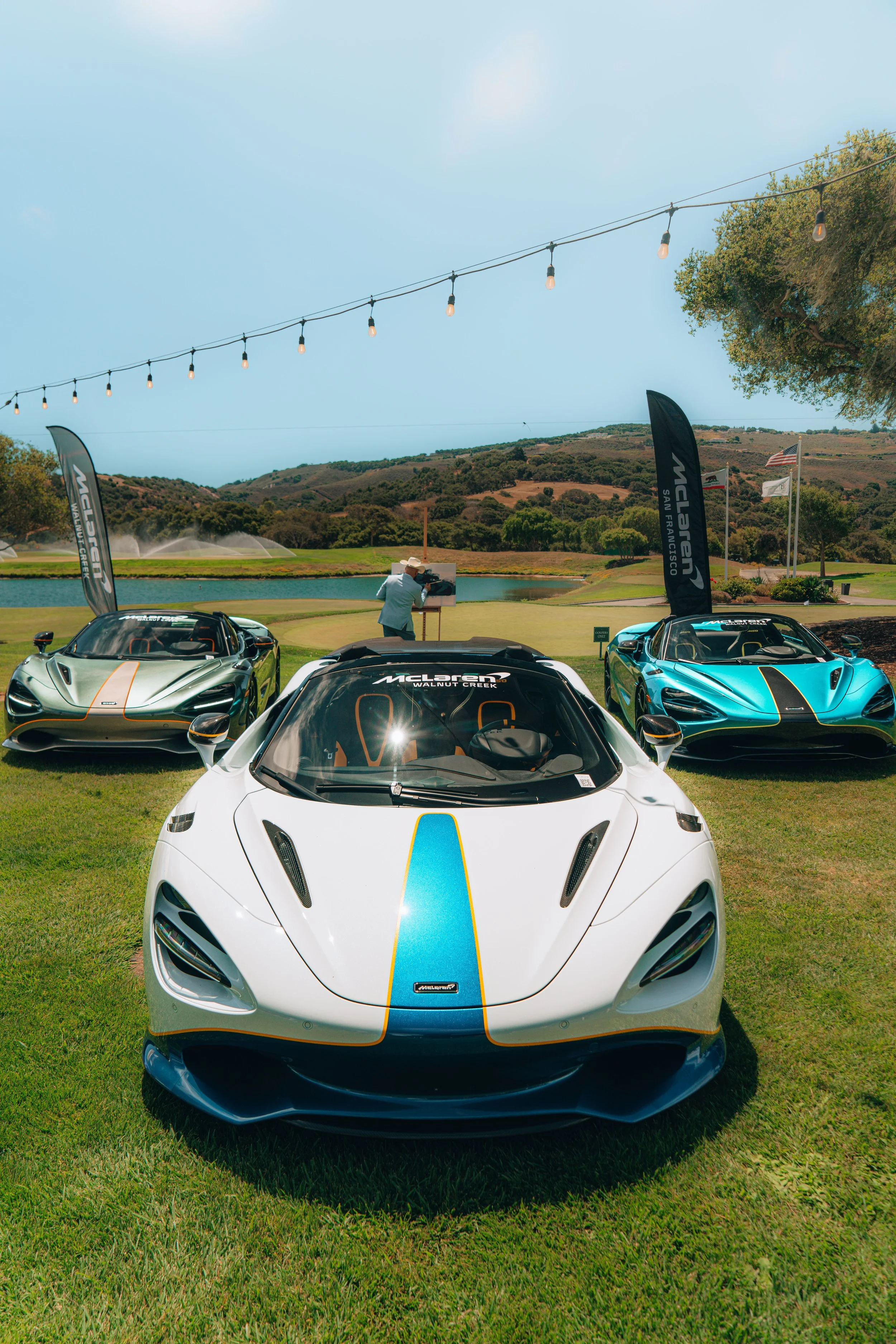 Three McLaren sports cars are displayed on a grassy area outdoors, with a lake, trees, and rolling hills in the background under a clear sky with string lights hanging overhead.