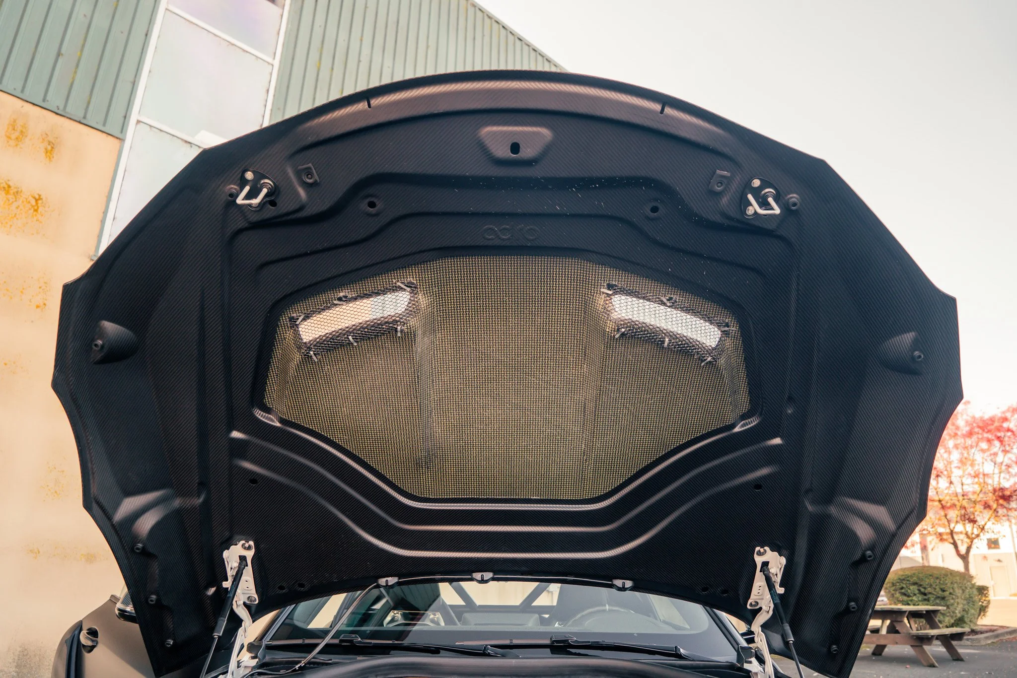 The hood of a car open, showing a carbon fiber lining and metallic components, against an industrial building background.