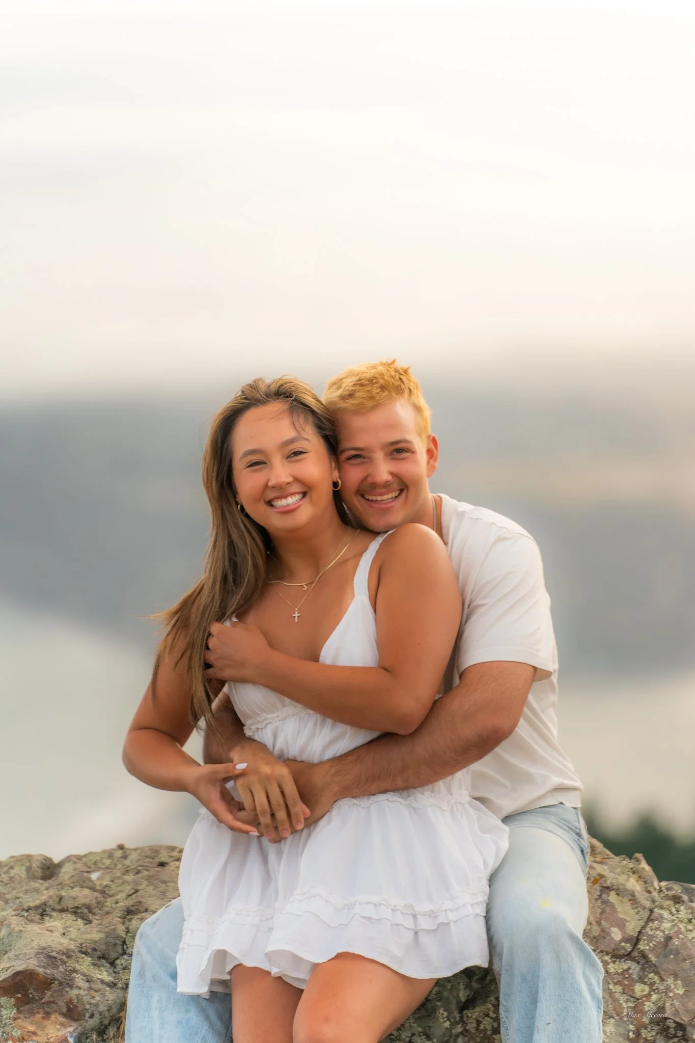 A happy couple sitting on a rock outdoors, embracing each other and smiling at the camera. The woman has long brown hair and wears a white dress, while the man has short blonde hair and wears a white shirt and light blue jeans. The background feature