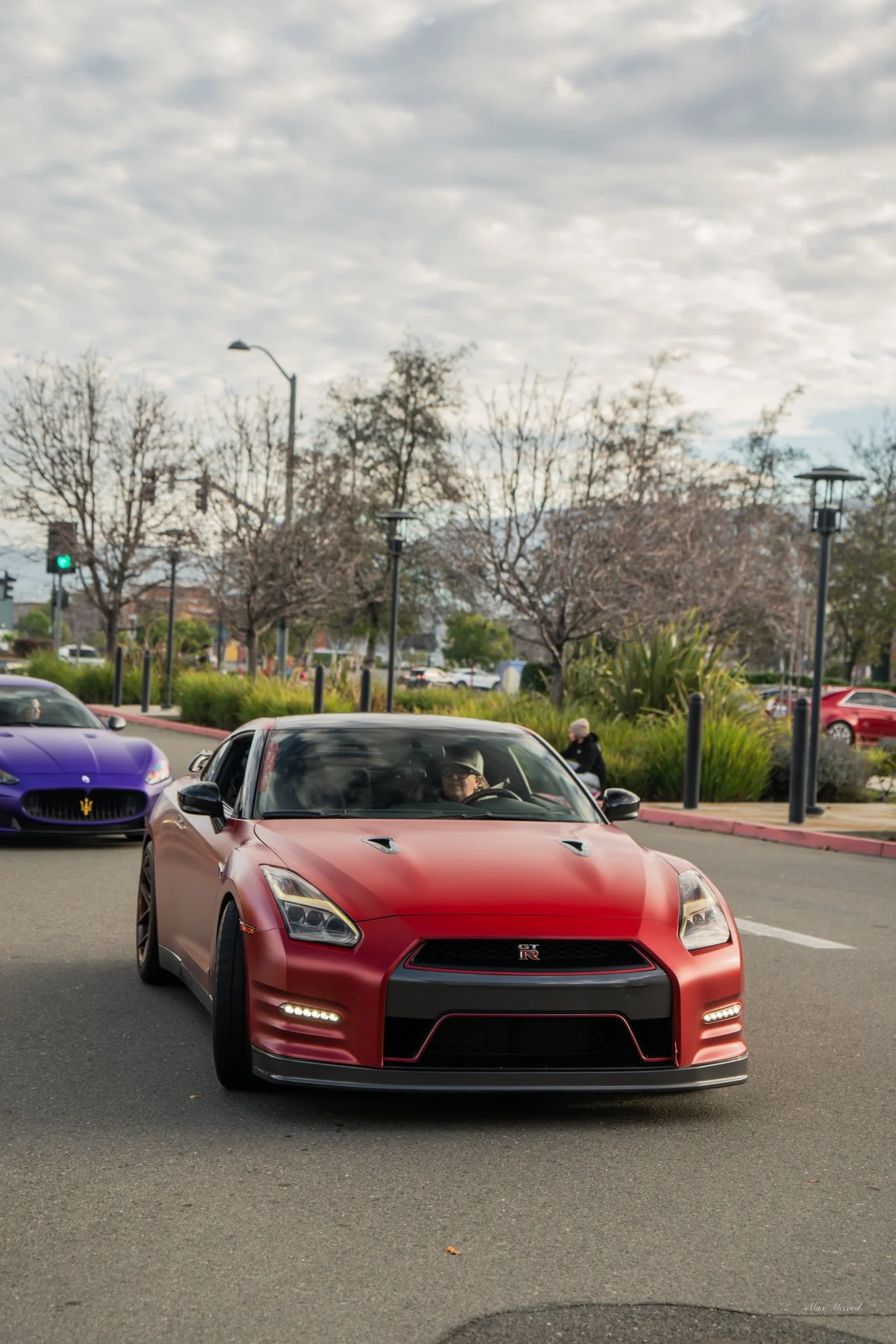A red Nissan GT-R sports car driving on a city street, with a purple Maserati behind it and trees with leafless branches in the background.