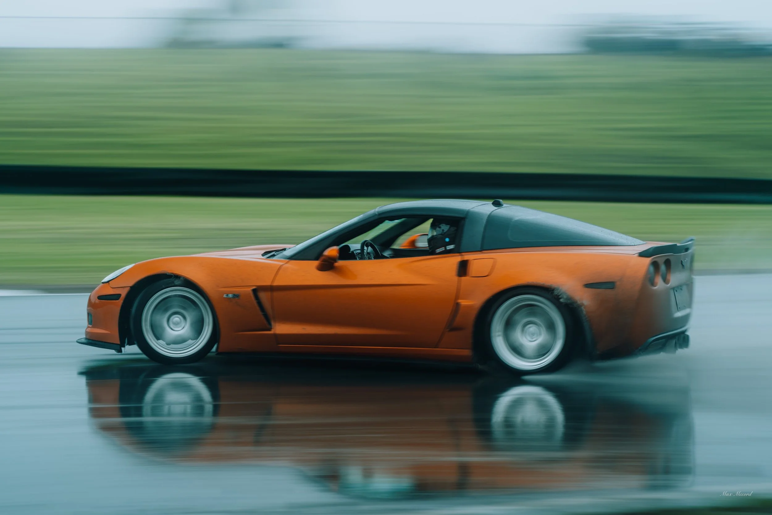 Orange sports car speeding on wet track with blurred green background and reflections on the surface.