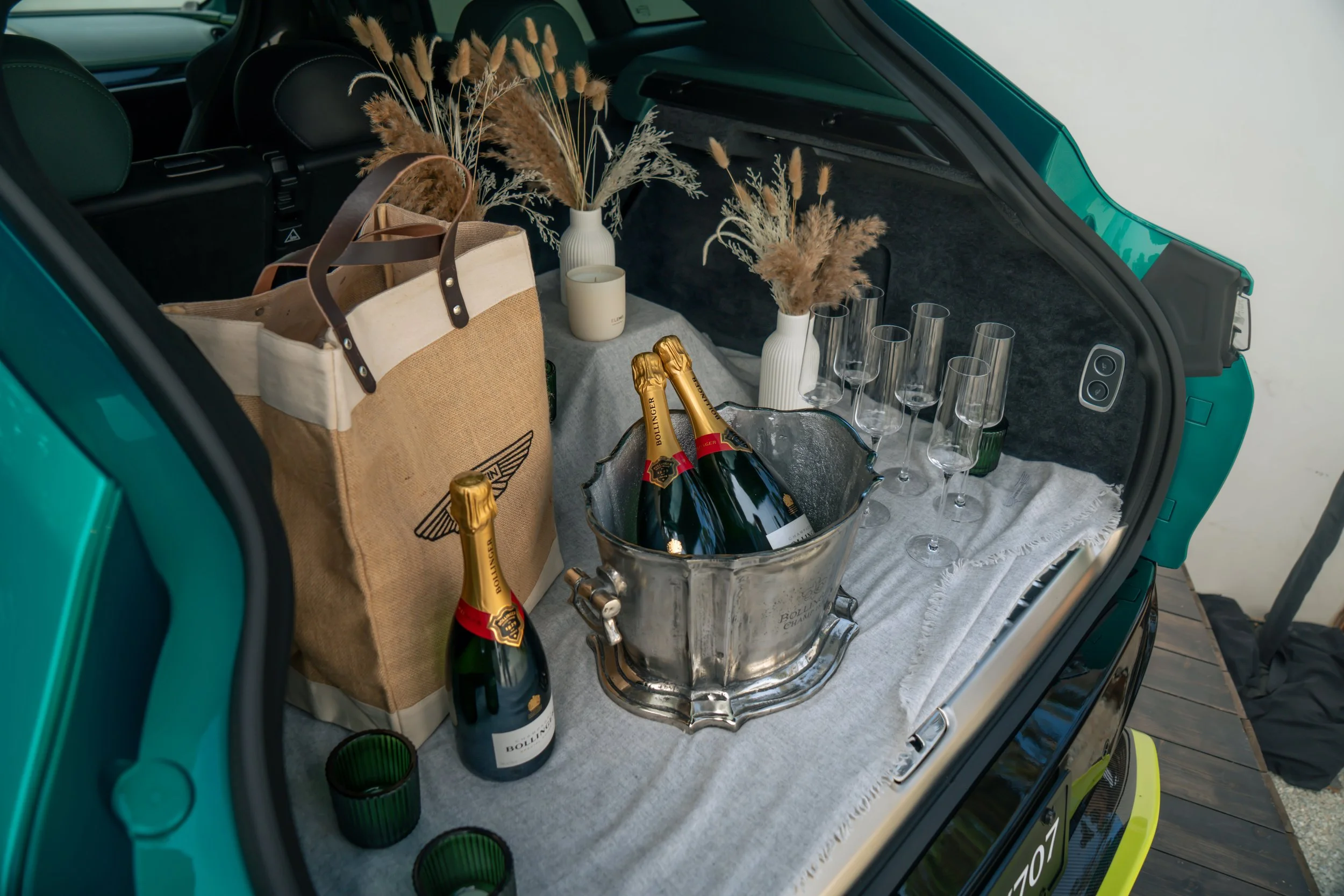The back of a car with a setup for a celebration, featuring bottles of champagne in an ice bucket, empty champagne glasses, decorative dried flowers in vases, paper bags, and a cloth covering the surface.