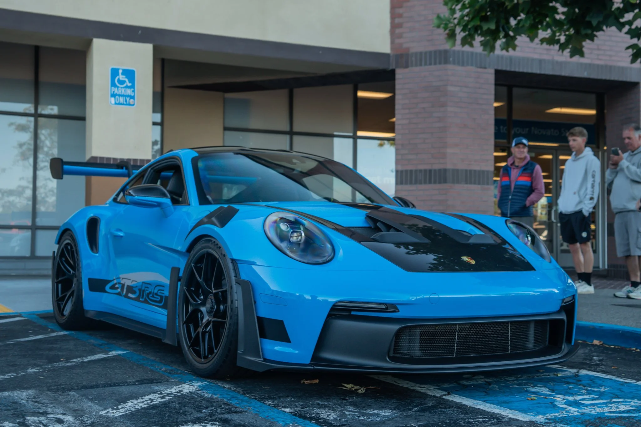 A blue Porsche racing car parked in a handicapped parking space outside a store, with three people standing nearby.