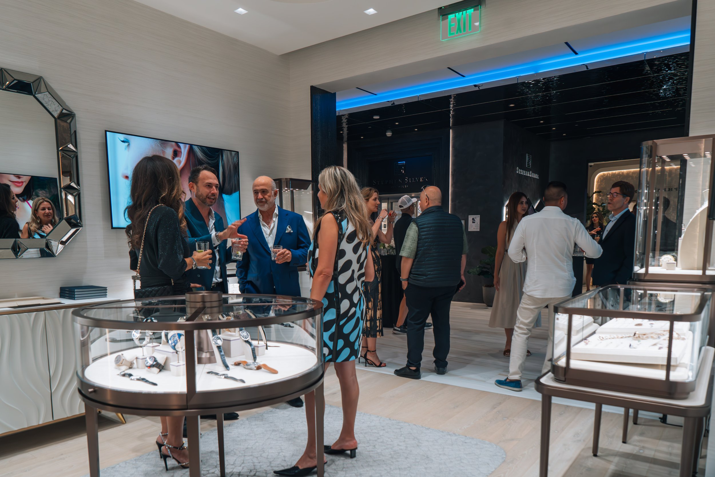 People socializing at a jewelry store or exhibit, displaying watches and accessories in glass cases, with a modern interior and a large TV screen in the background.