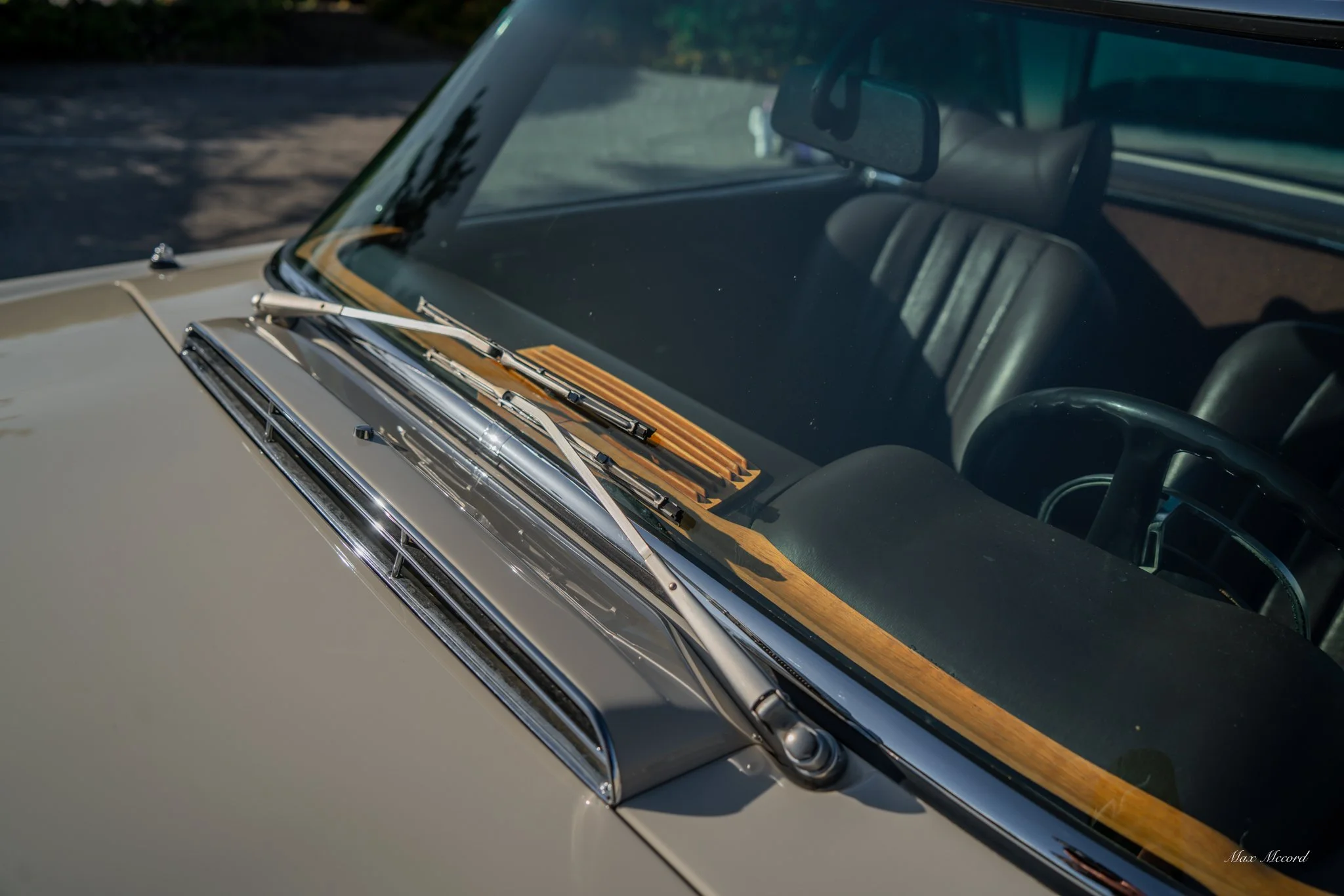 Close-up of the front windshield of a vintage beige car, showing wipers, dashboard, and interior seats, with sunlight reflecting off the chrome details.