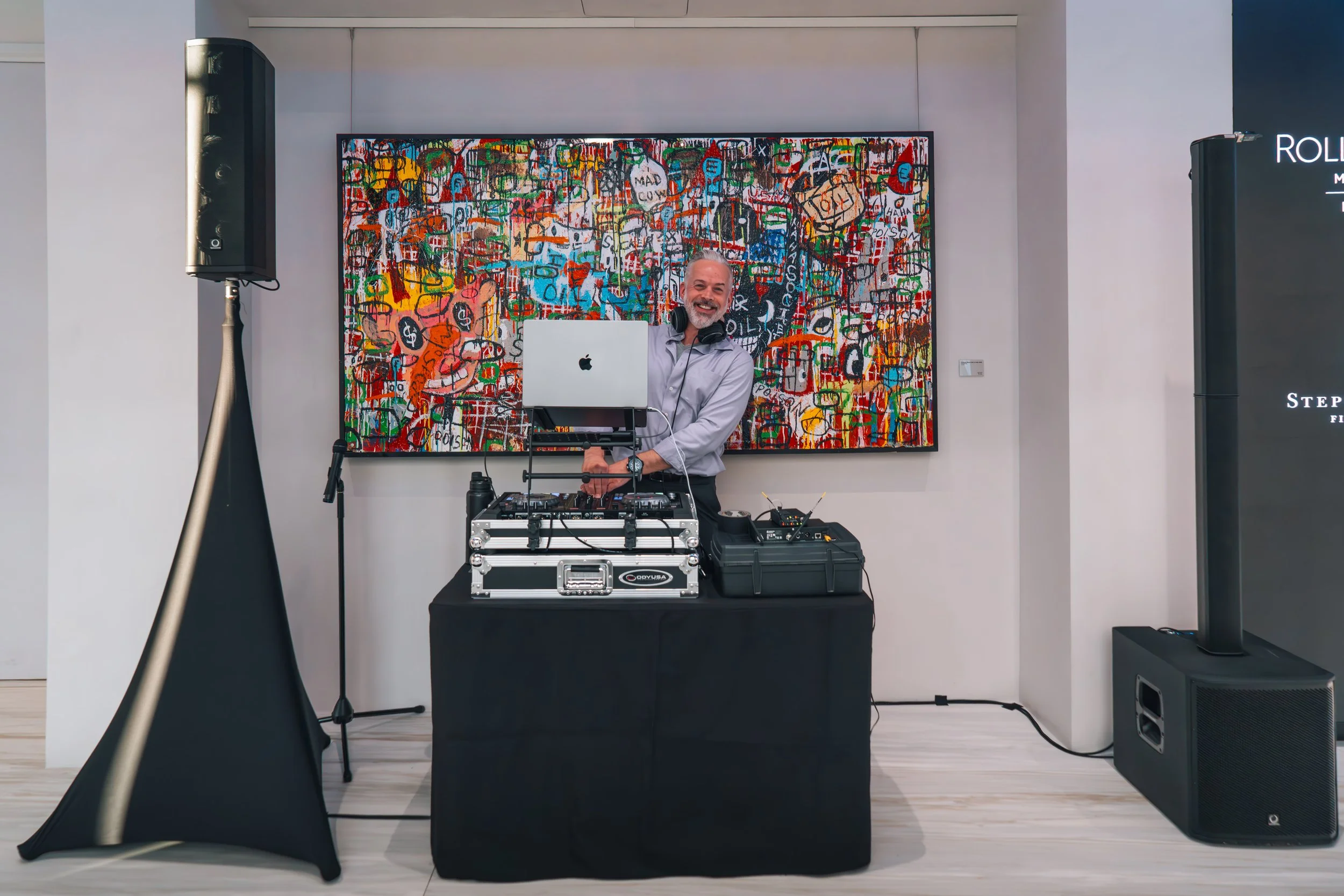 A smiling DJ with headphones around his neck, standing behind a DJ console with a laptop, in an art gallery, with colorful abstract artwork on the wall and large speakers on either side.