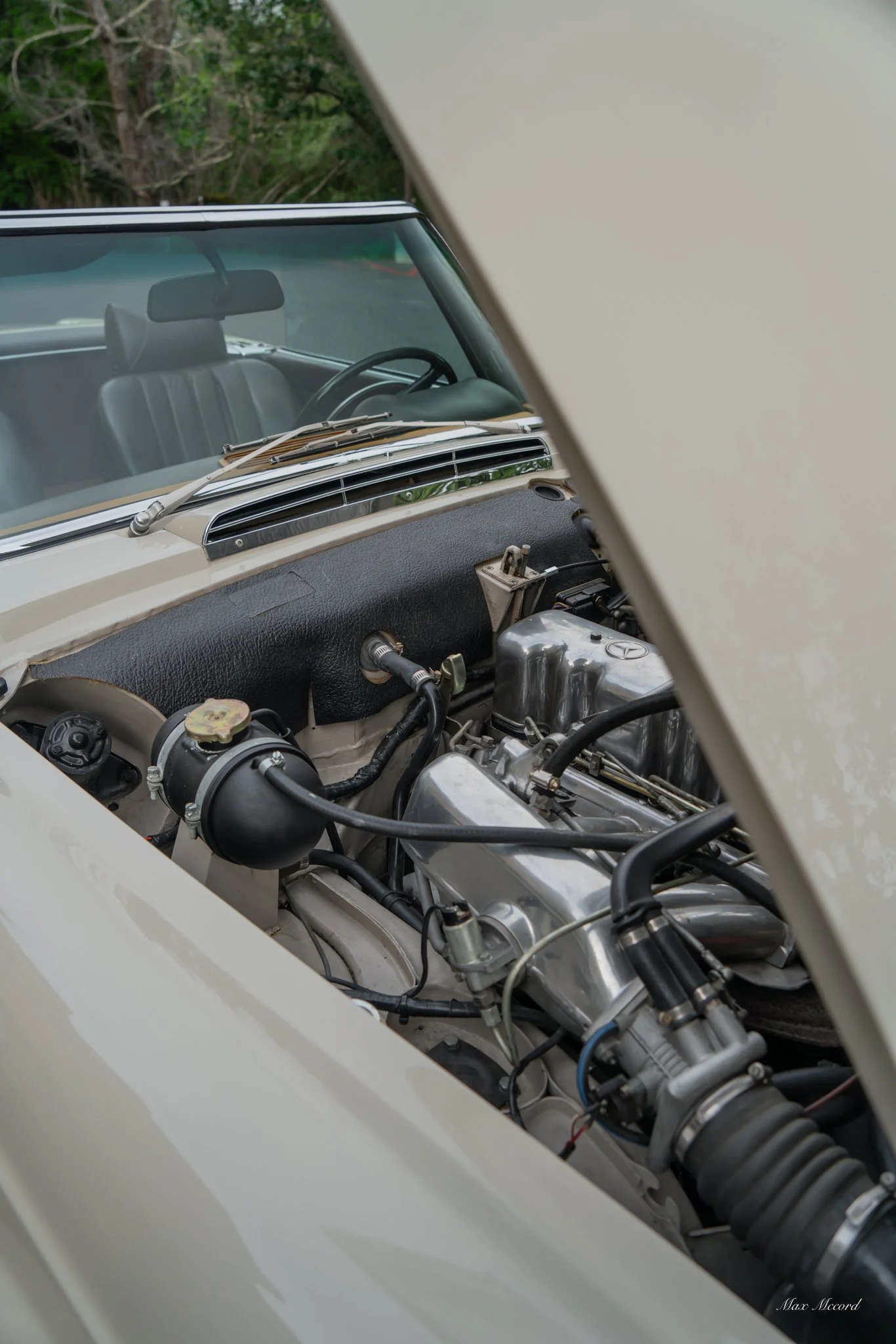 Close-up of a classic car engine viewed through an open hood, with the windshield and interior visible in the background.
