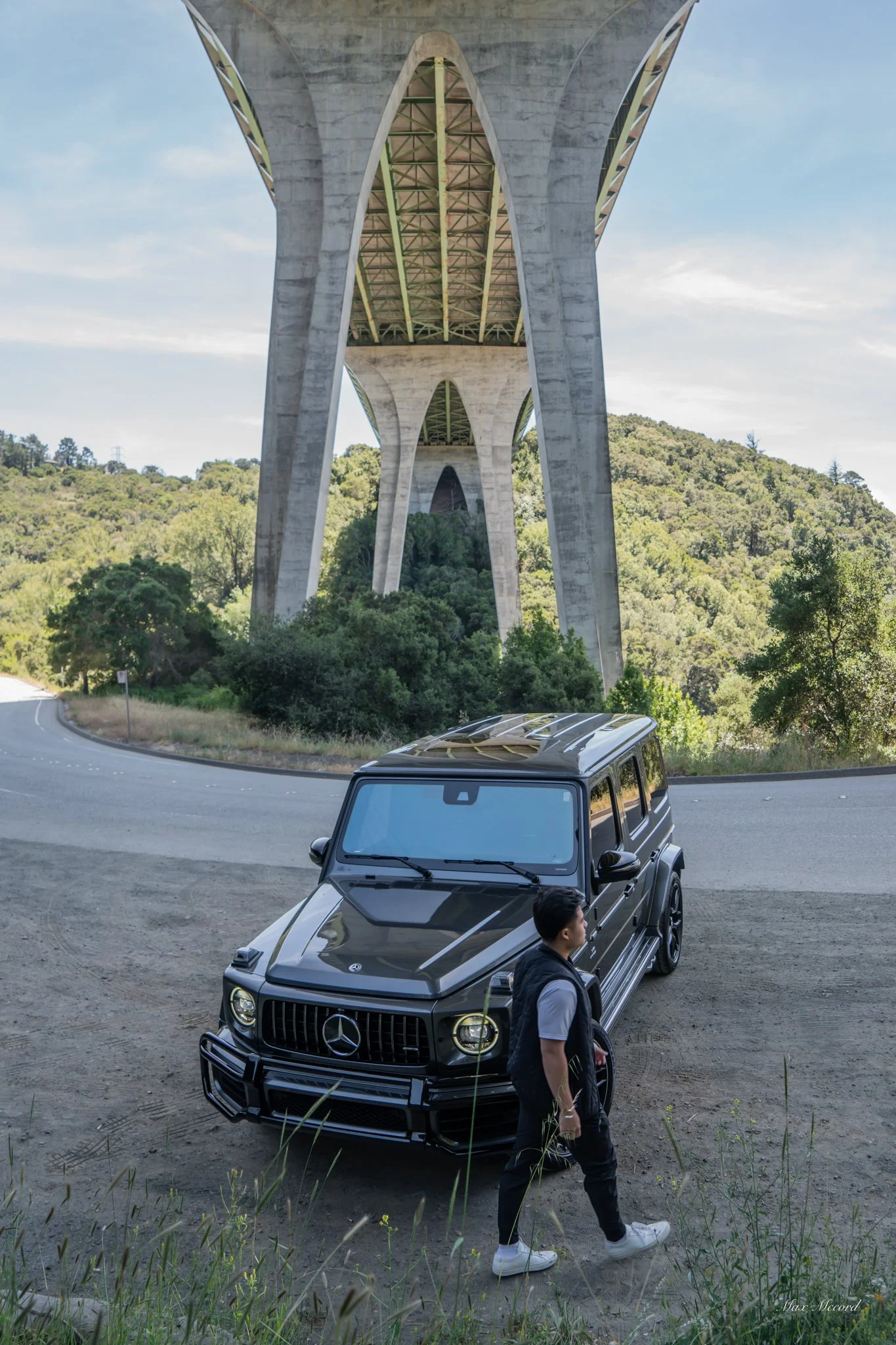 A man walking past a black Mercedes-Benz G-Class SUV parked under a large concrete bridge over a curved road in a rural area with trees and hills in the background.