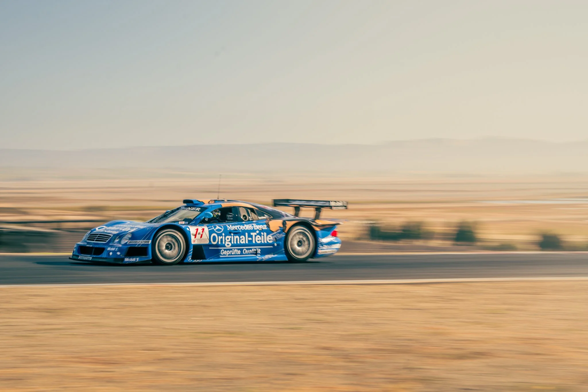 A blue Mercedes-Benz race car moving at high speed on a racetrack in a desert landscape during daytime.