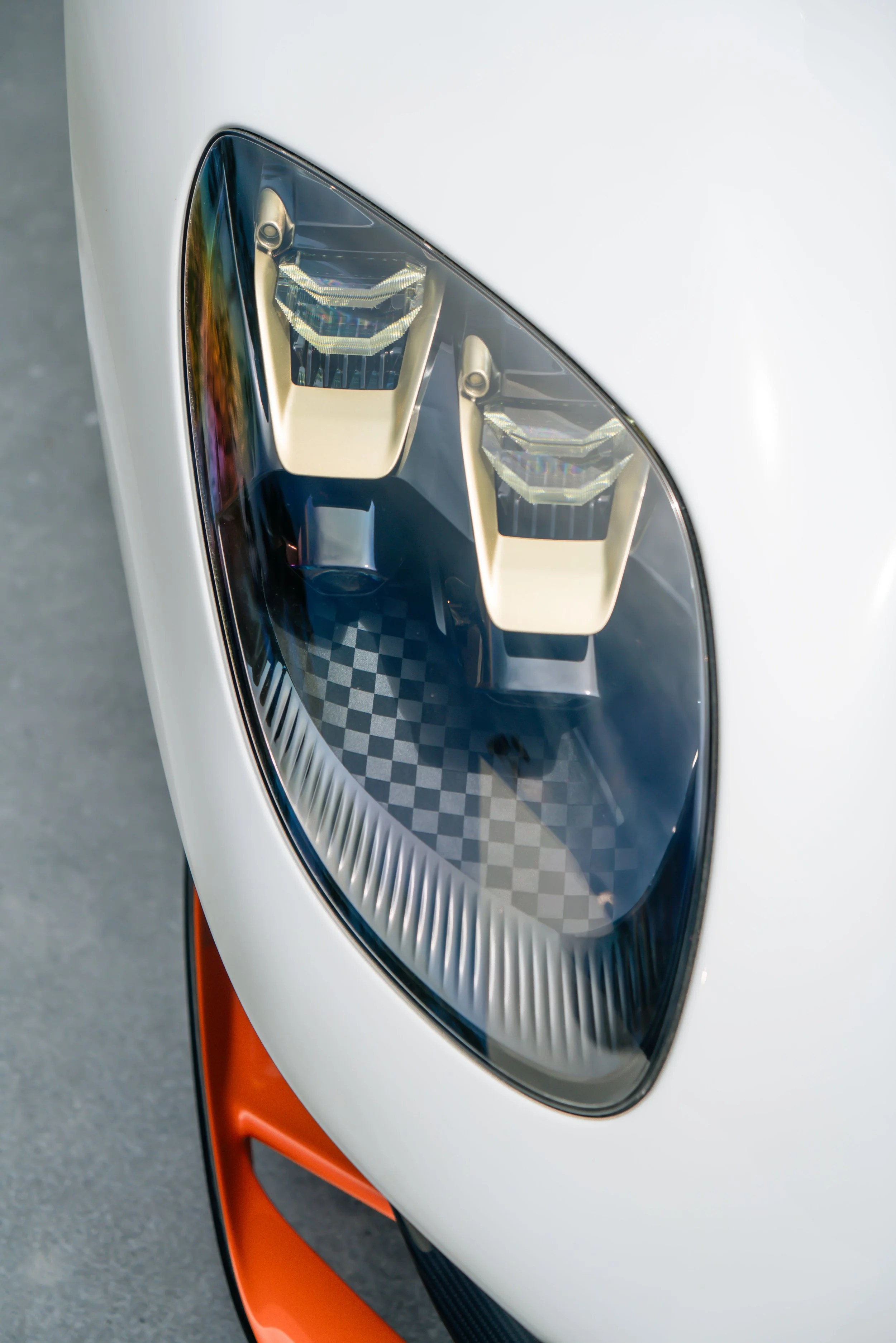 Close-up of a sports car headlight with a clear cover, saving a white and orange body.