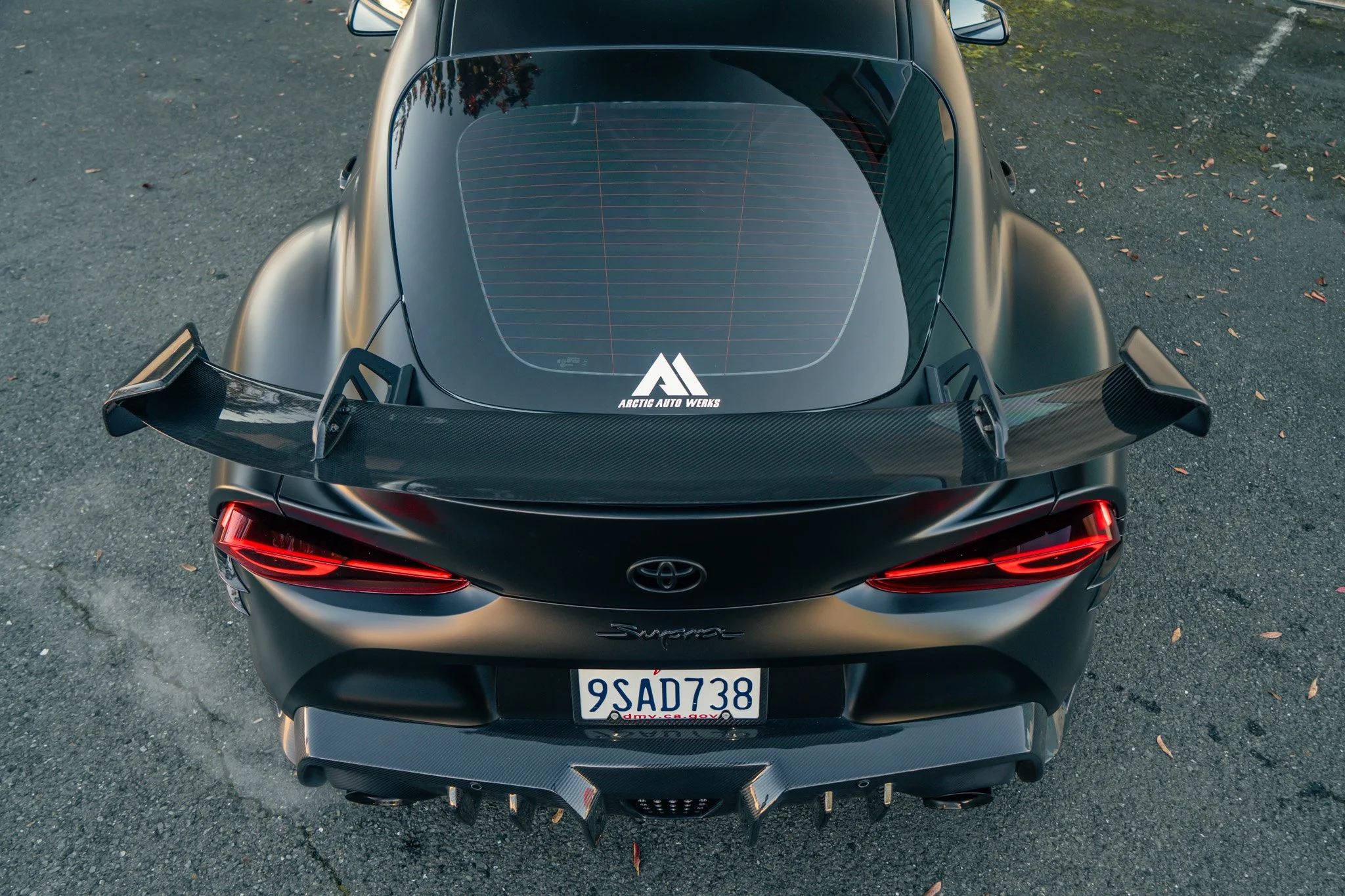 Top-down view of a black Toyota Supra sports car with a large rear spoiler, a carbon fiber diffuser, and a decal that reads 'Arctic Auto Werks' on the rear window, parked on an asphalt surface.