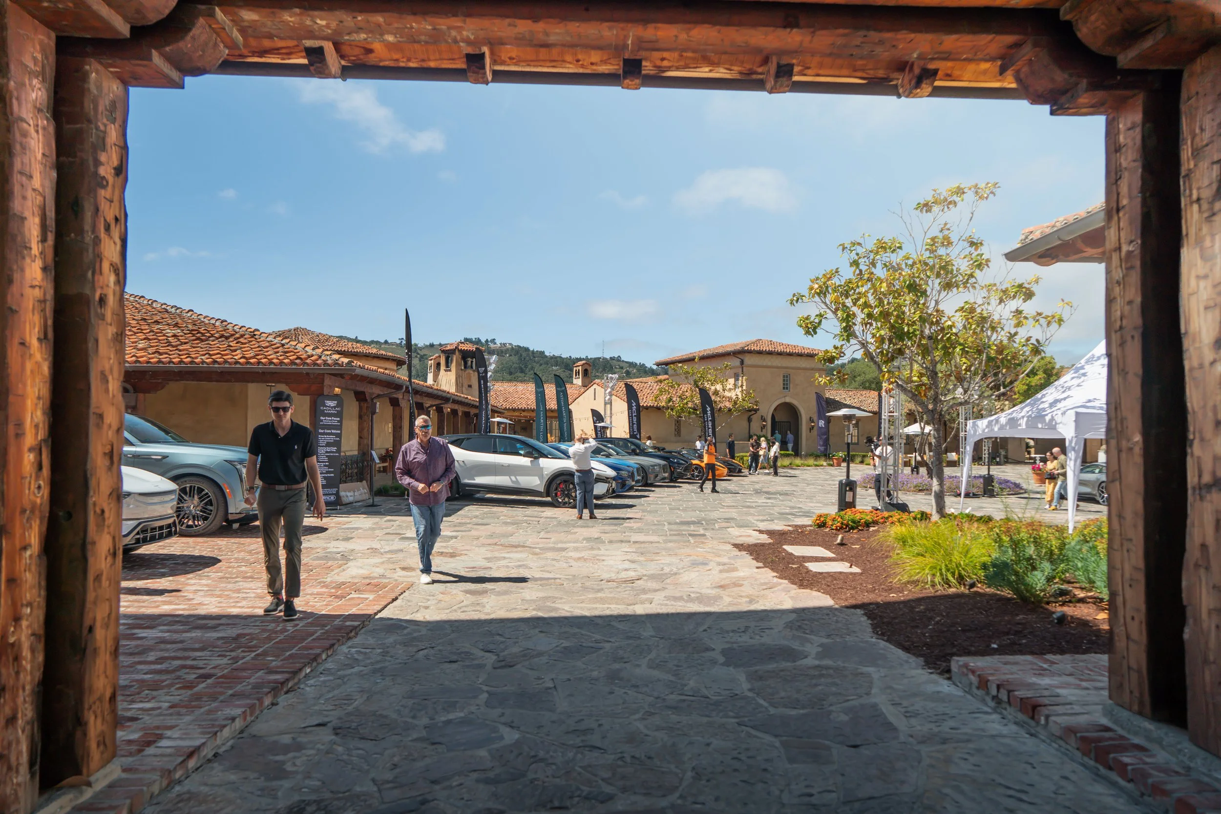 View of a modern outdoor car exhibition from a wooden archway, with luxury cars on display, people walking around, and a sunny sky with a few clouds.