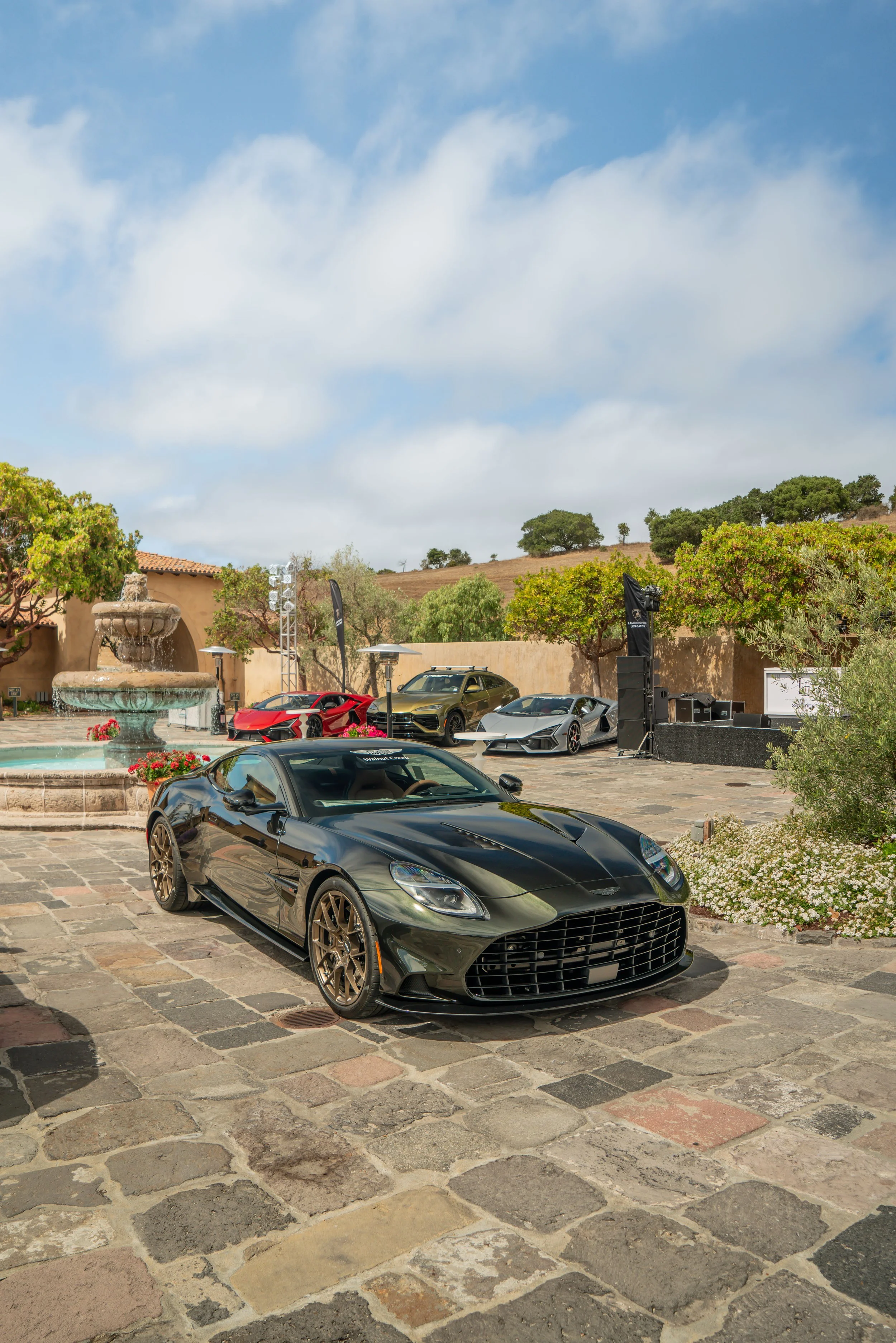Black luxury car parked on a stone driveway with several high-end sports cars and an outdoor speaker setup in the background, surrounded by trees and a fountain.