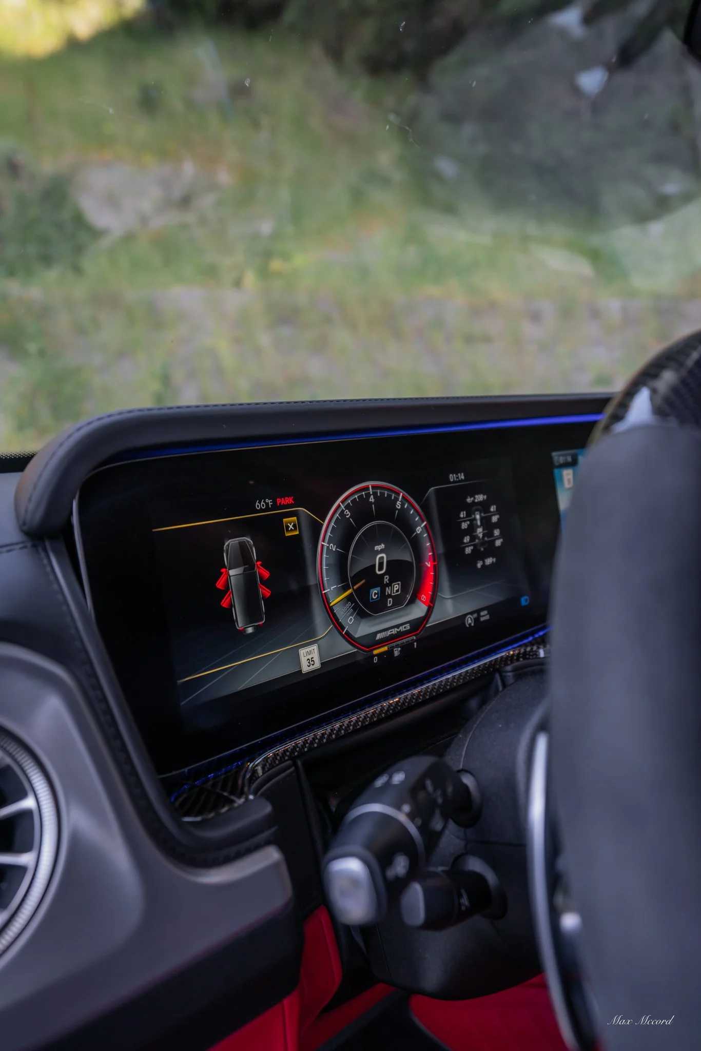 Digital dashboard of a car showing the speedometer at zero, the gear in park, and warning indicators. The background reveals a natural outdoor scene with grass and rocks through the windshield.
