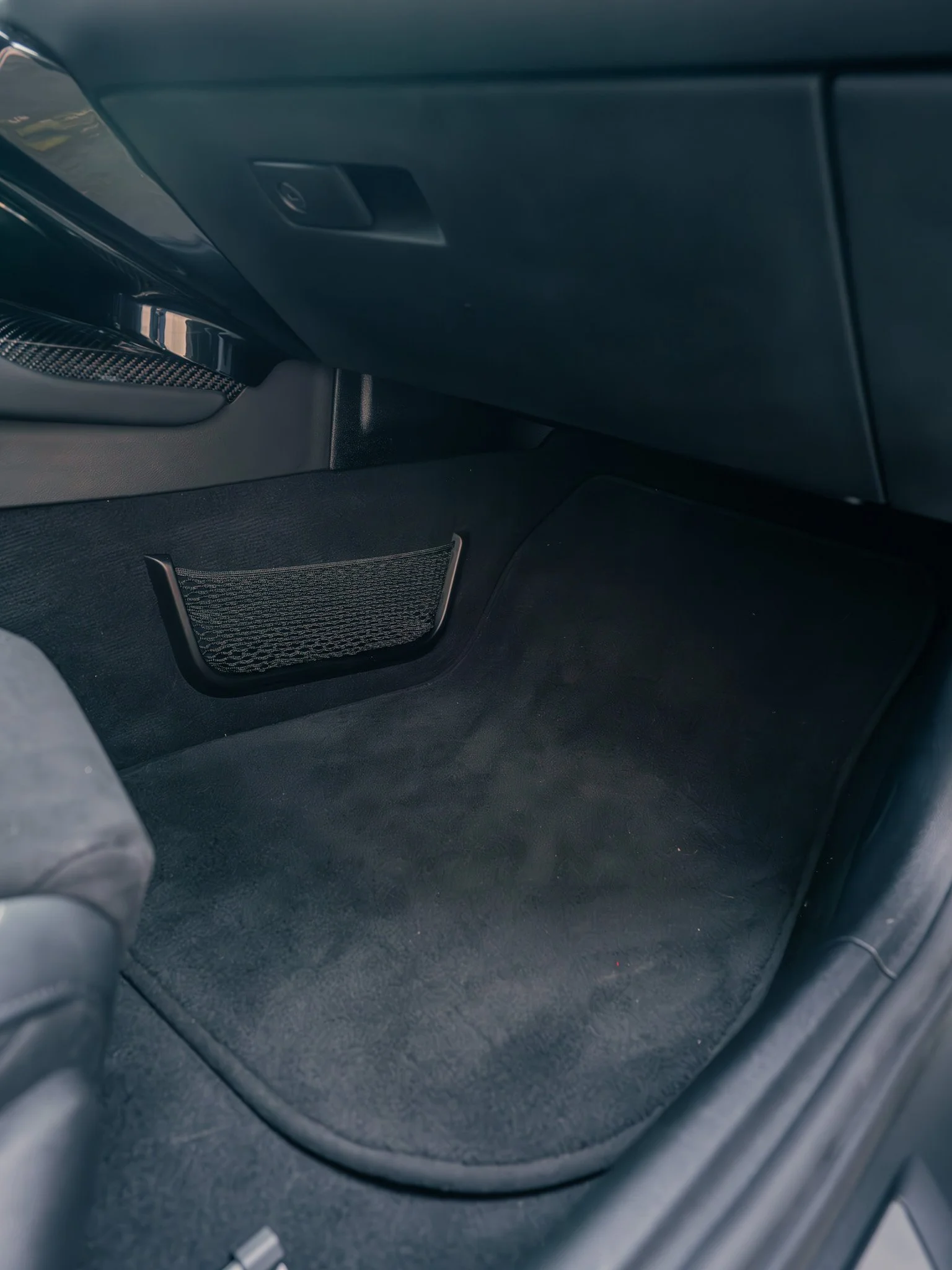 Photo of the front passenger side footwell of a car, showing a black floor mat, a storage net, and part of the dashboard.