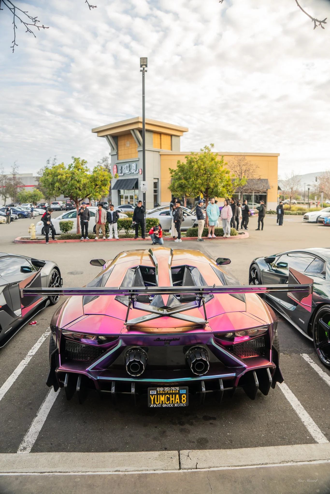 Rear view of a vivid purple and black luxury sports car, parked in a lot with other cars, behind a group of people standing outside a restaurant in a commercial area.