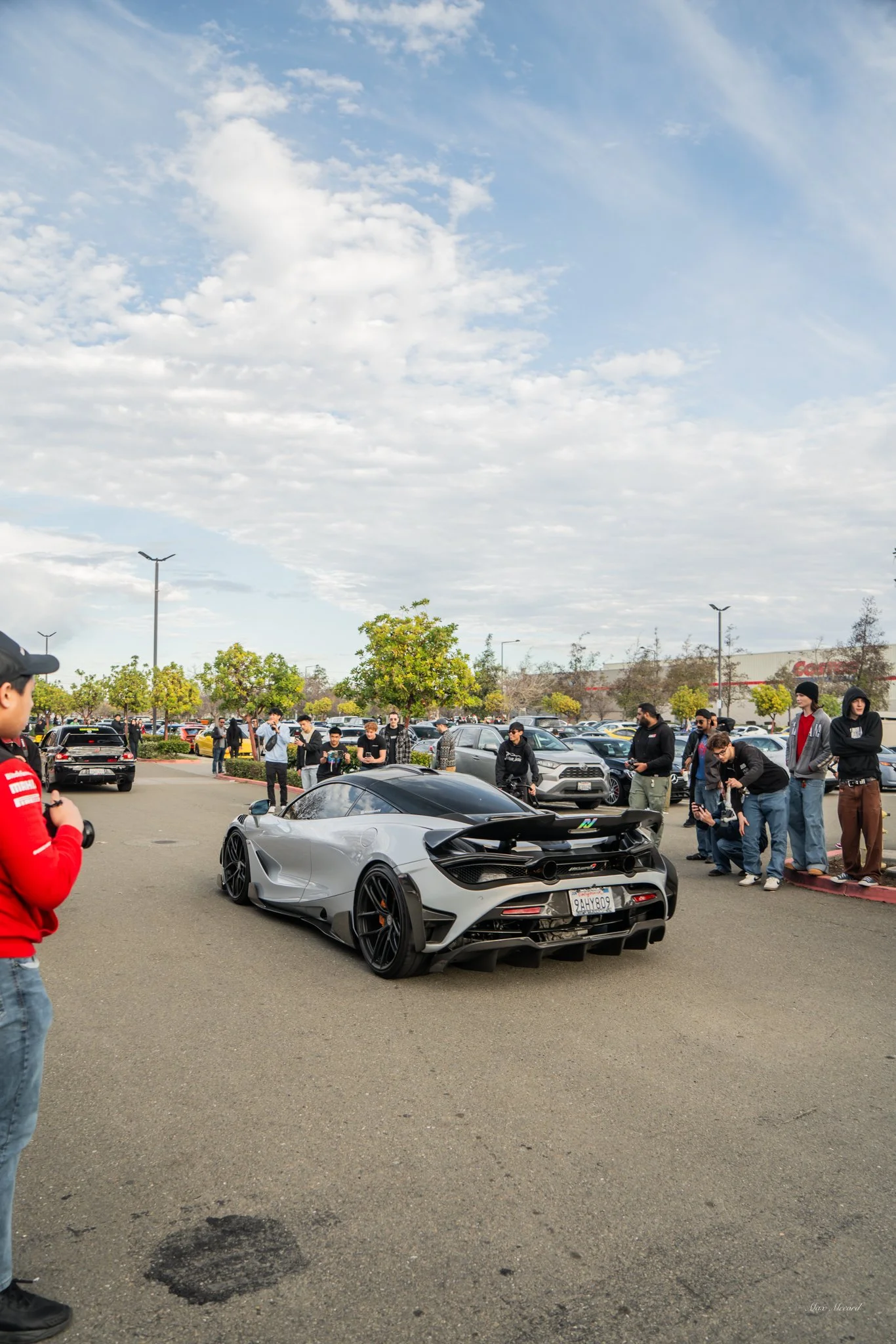 A silver McLaren sports car parked in a parking lot with a group of people gathered around, some taking photos. The sky is partly cloudy with scattered clouds, and there are trees and other cars in the background.