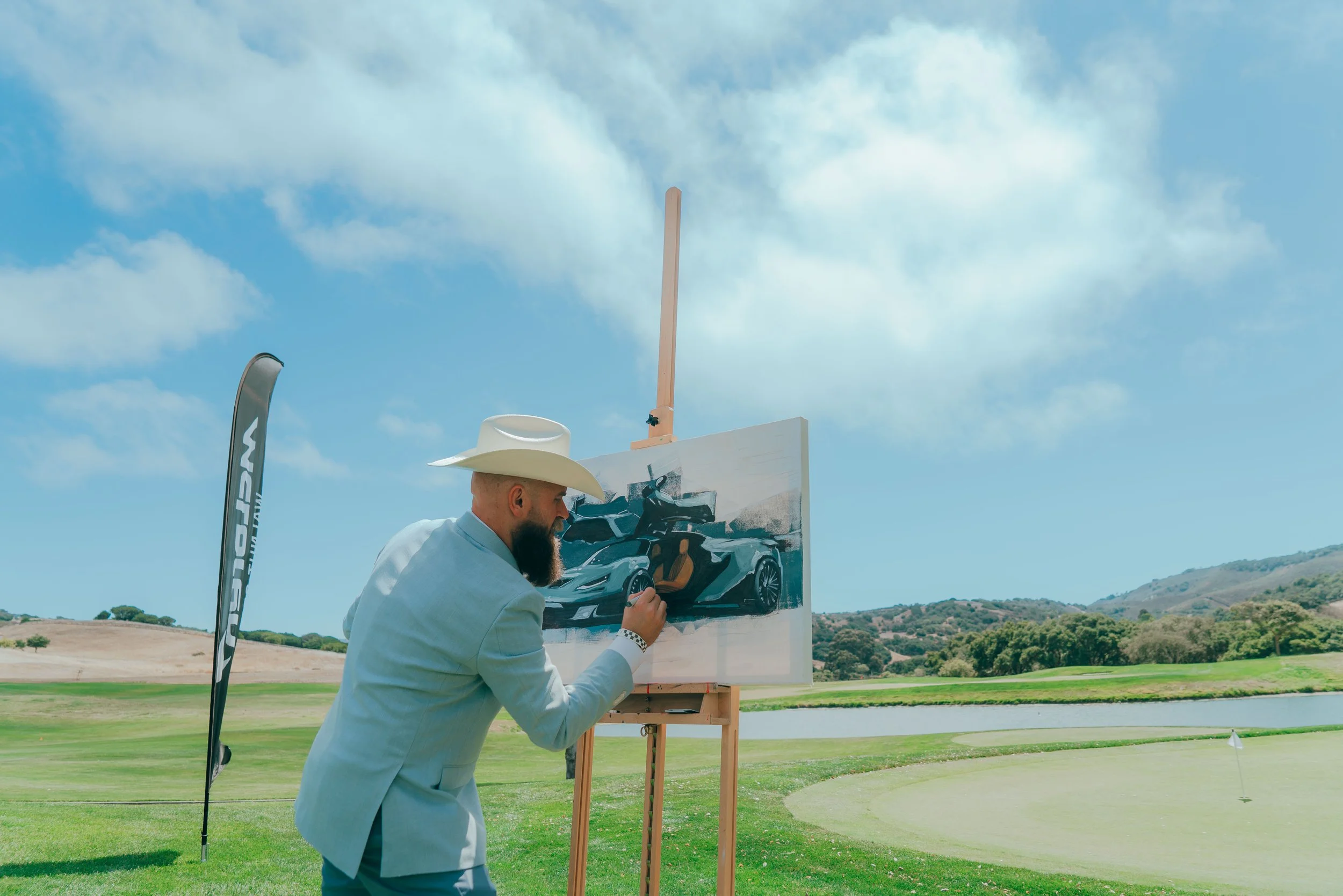 Man in a light blue blazer and cowboy hat painting a sports car on a canvas outdoors, with golf course and hills in the background.