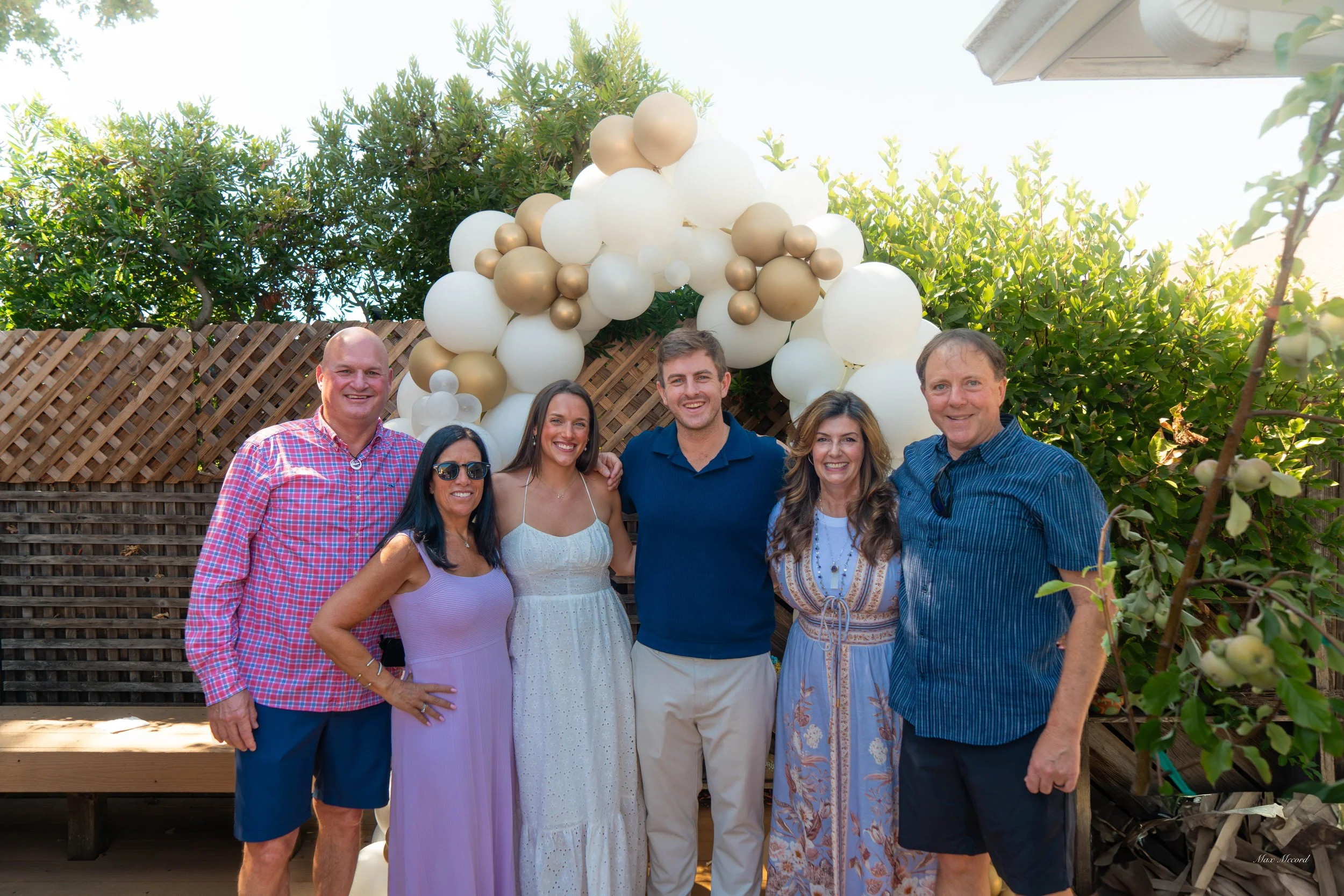 Group of six people standing together in front of a balloon arch, outdoors in a backyard on a sunny day.