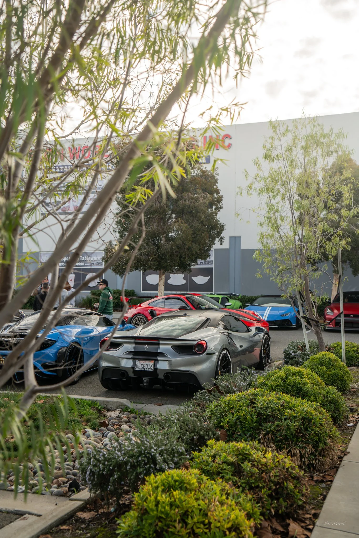Multiple exotic sports cars parked in an outdoor lot, with some greenery and trees in the foreground, and a building with signage in the background.