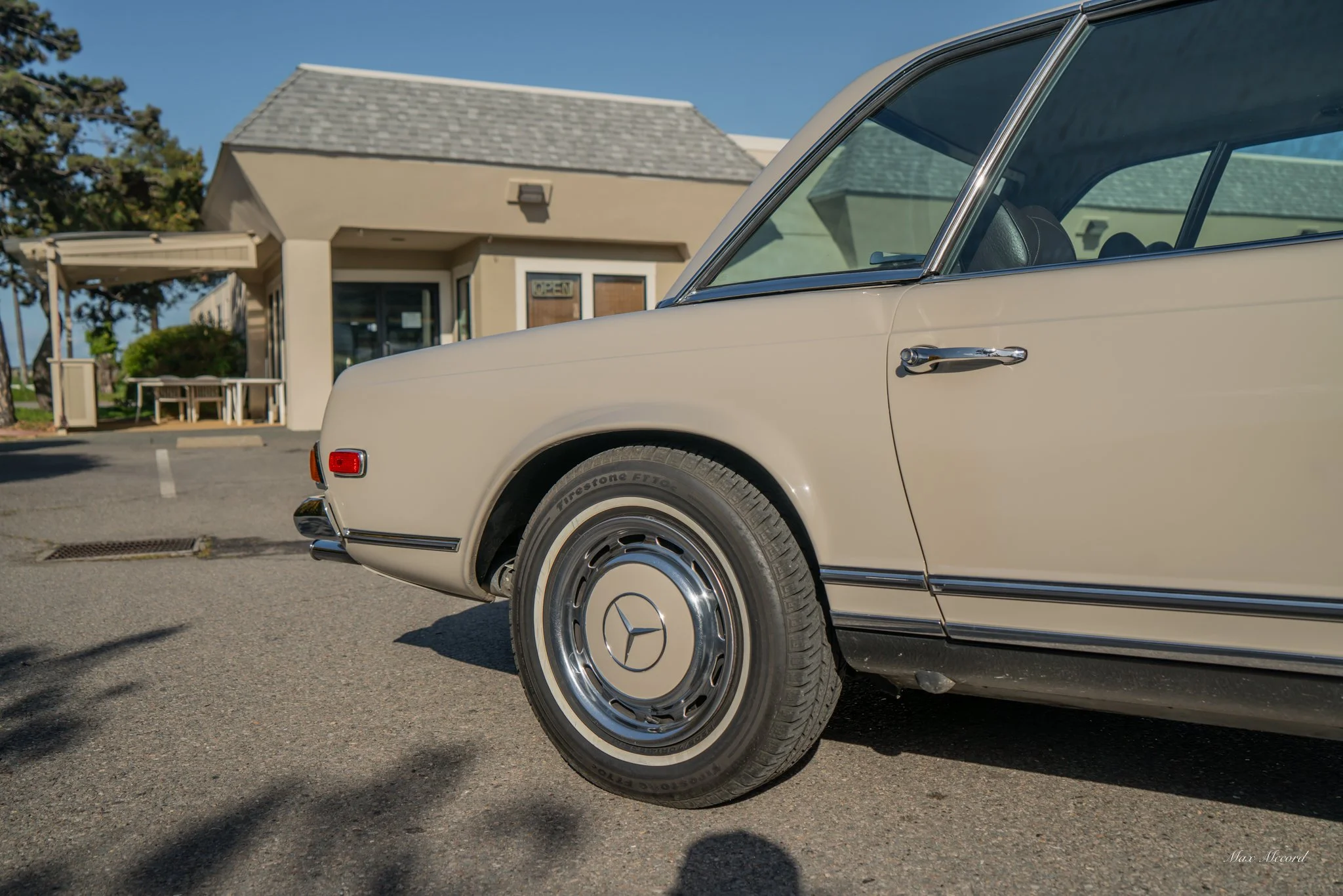 Close-up of the front part of a cream-colored vintage Mercedes-Benz car parked on a street in front of a building with a gray roof and beige exterior.