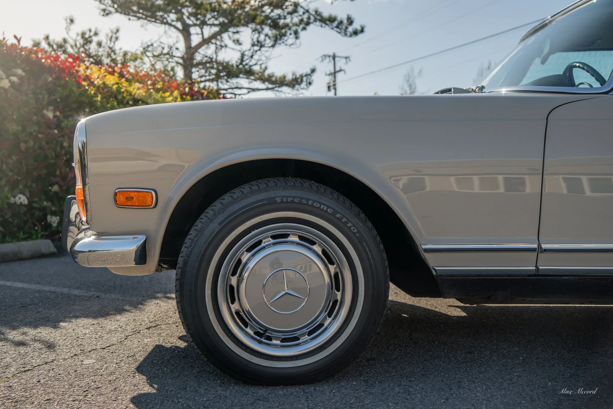Close-up of the front part of a vintage silver Mercedes-Benz car, focusing on the wheel and tire, with a Mercedes logo on the hubcap, parked on a paved road near some bushes with red, yellow, and white flowers, and trees in the background.