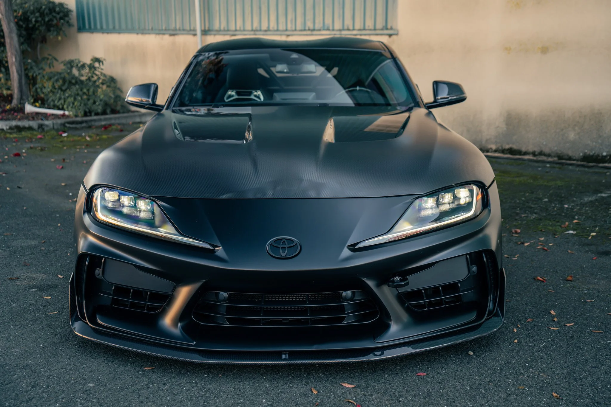 Front view of a black Toyota sports car with sleek design, LED headlights, and carbon fiber hood parked on asphalt ground near a wall.