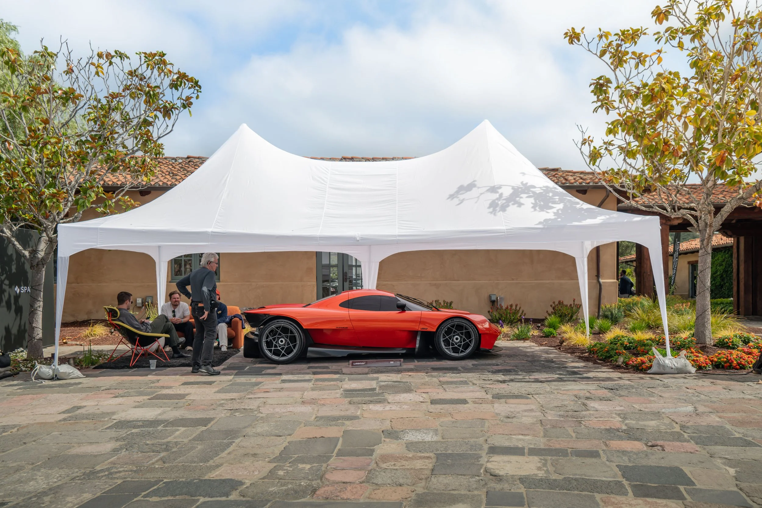Red sports car under a white outdoor tent with people sitting and standing nearby in a courtyard with trees and buildings.