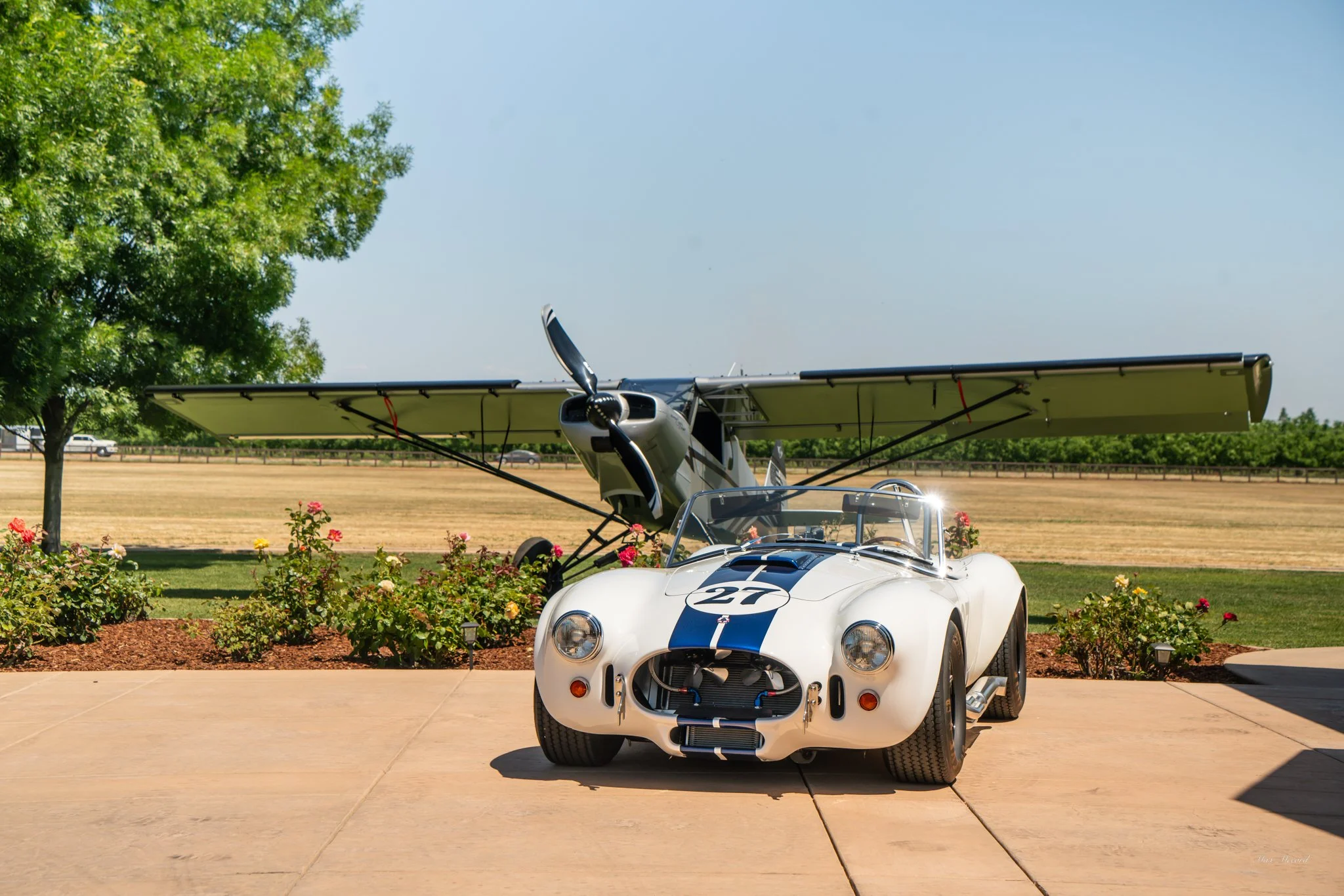A vintage race car with the number 27 on it parked in front of a small propeller airplane at a sunny outdoor location, with trees and a field in the background.
